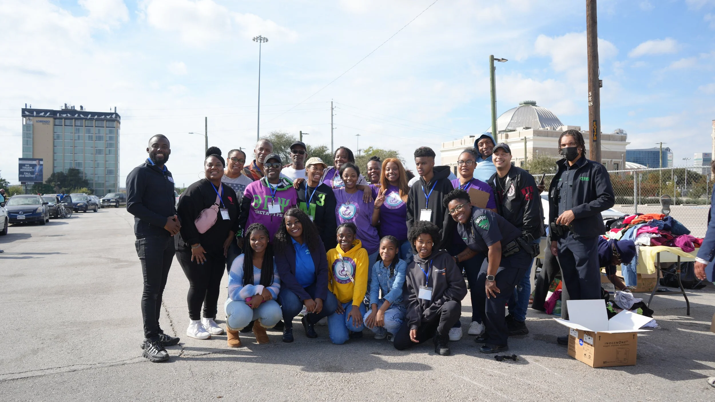 Group of diverse people, including children and adults, posing outdoors on a sunny day with a cityscape background, some wearing purple shirts and others in casual or police uniforms, standing in front of a table with clothes and a cardboard box.
