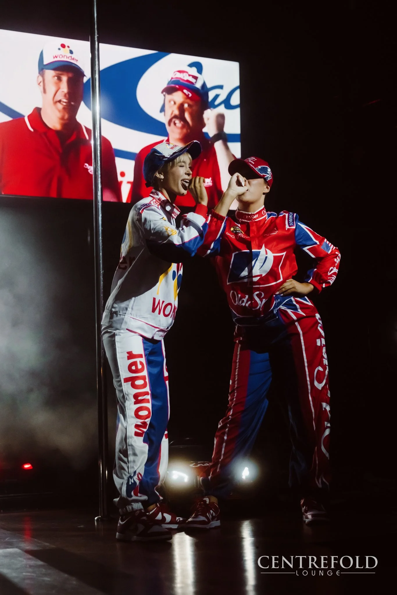 Two young race car drivers, a girl and a boy, celebrating on stage in racing suits and caps. The girl is smiling and touching the boy's face as he stands confidently with his hand on his hip. There is a screen behind showing two men in red uniforms a