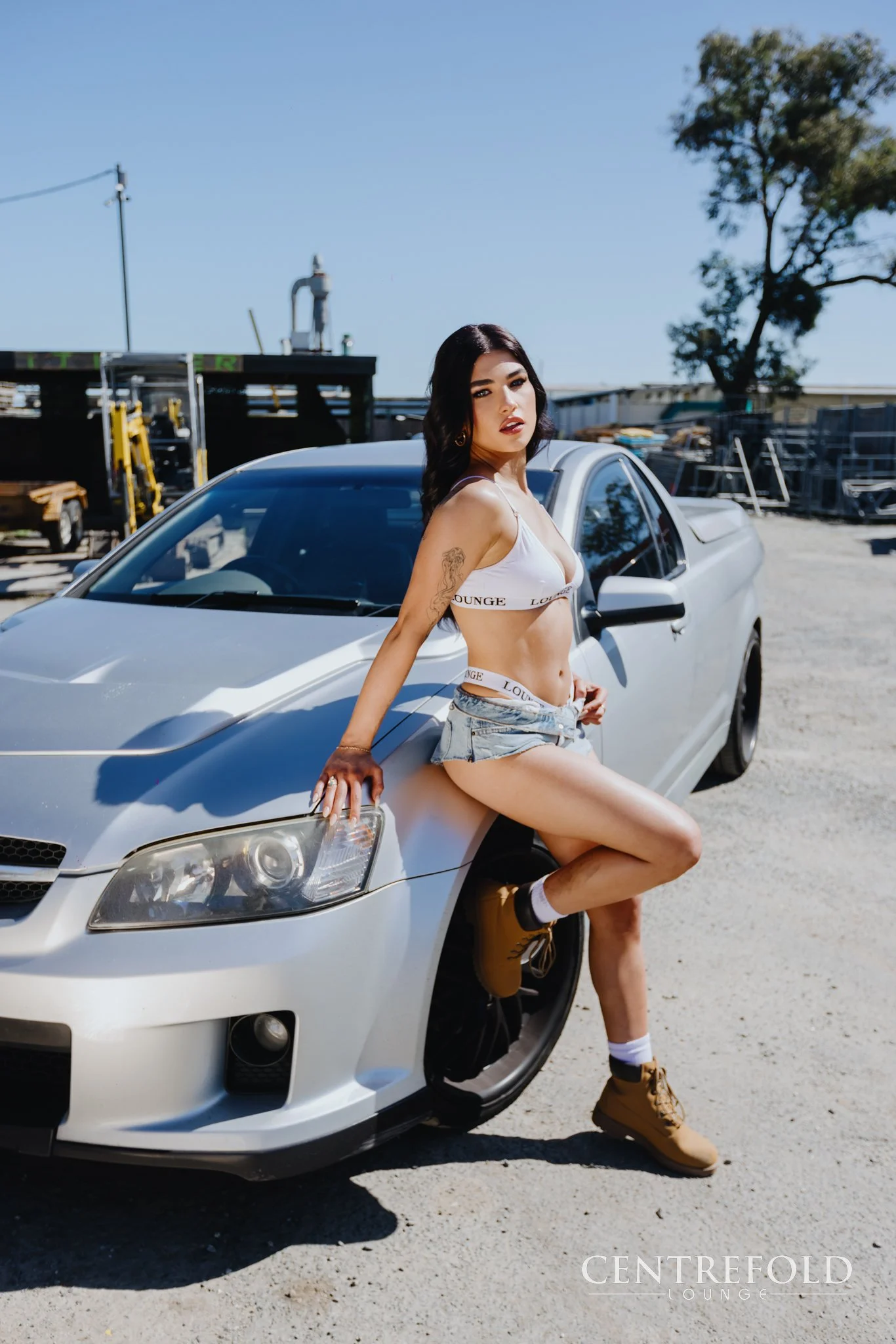 A woman with dark hair, wearing a white sports bra, denim shorts, and tan boots, posed leaning against a silver car in an outdoor setting on a sunny day.