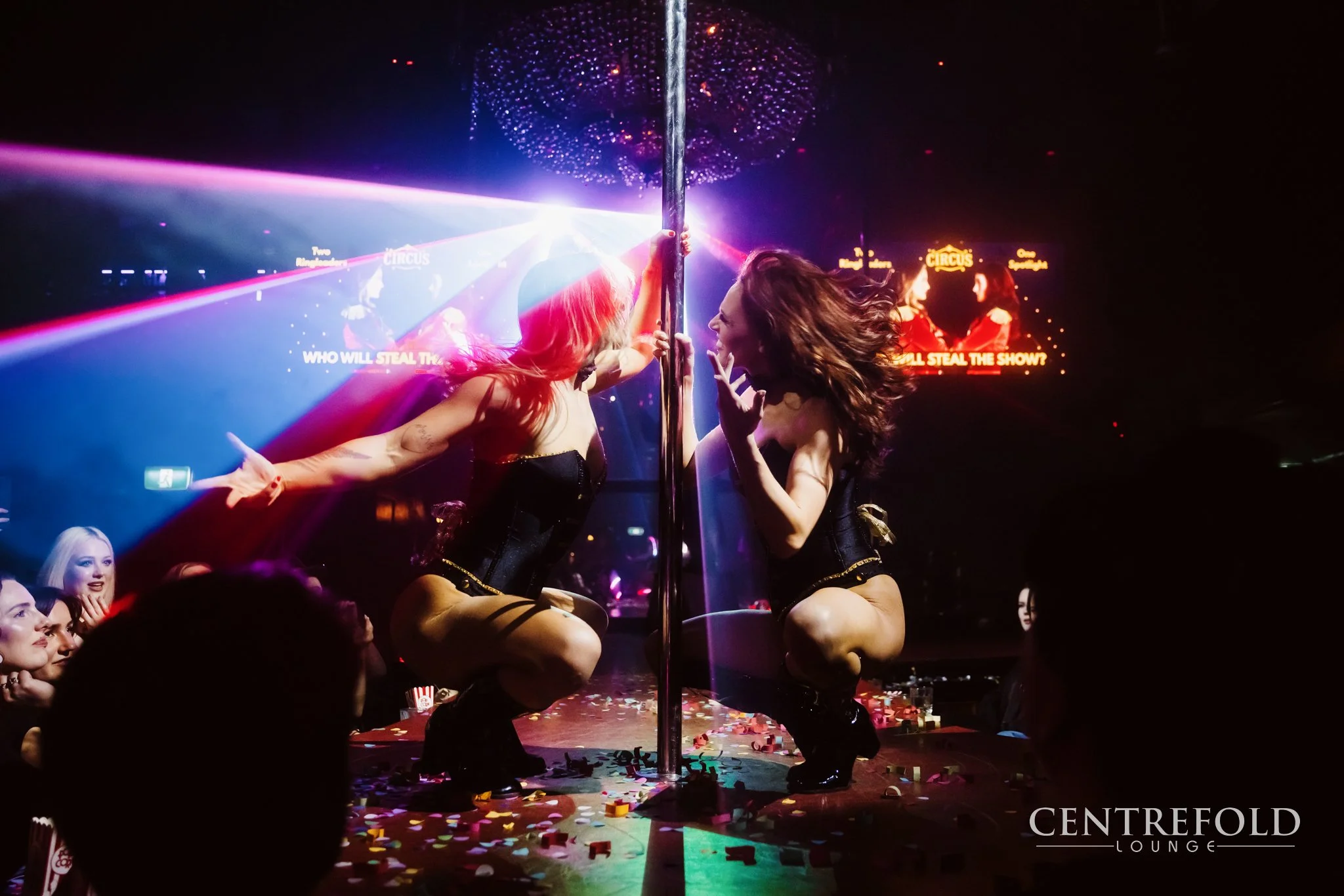 Two women performing a sensual dance on a stage with a pole, surrounded by colorful lights and an audience, at Centrefold Lounge.