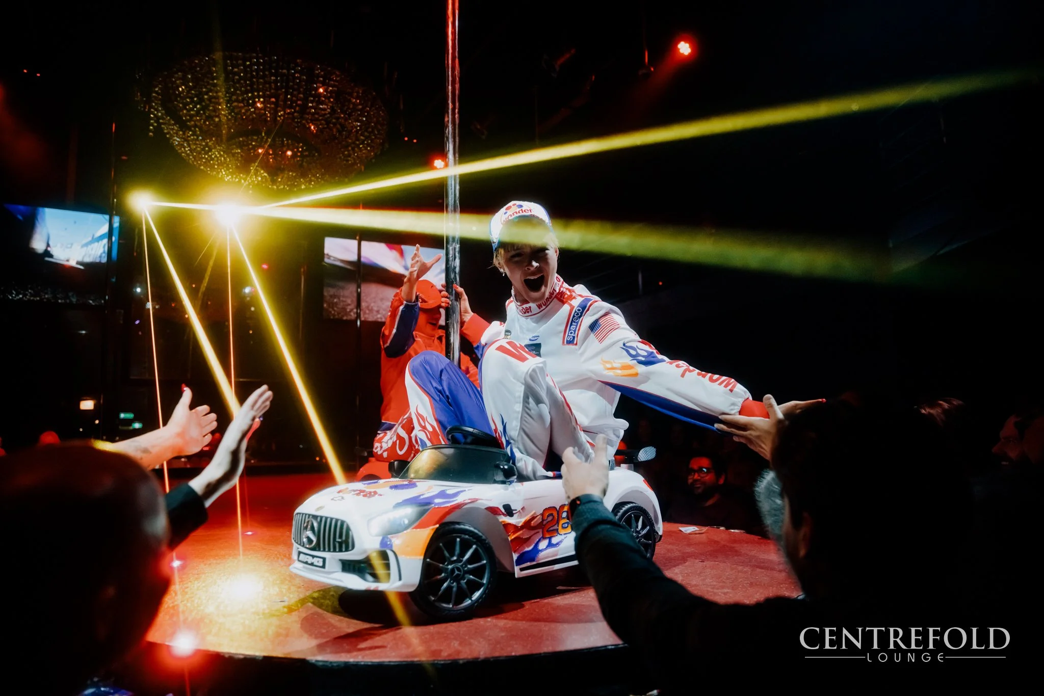 A woman in racing gear sitting in a toy car, celebrating on stage while people reach out to her at a party with colorful lighting and a chandelier.