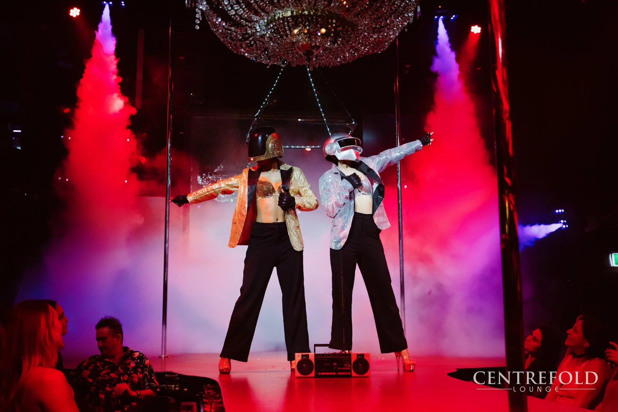 Two performers in shiny jackets and high heels stand on stage wearing helmets. They are near a pole, with one pointing and the other holding a microphone, surrounded by colorful fog and lighting. Audience members are sitting at tables in the foregrou
