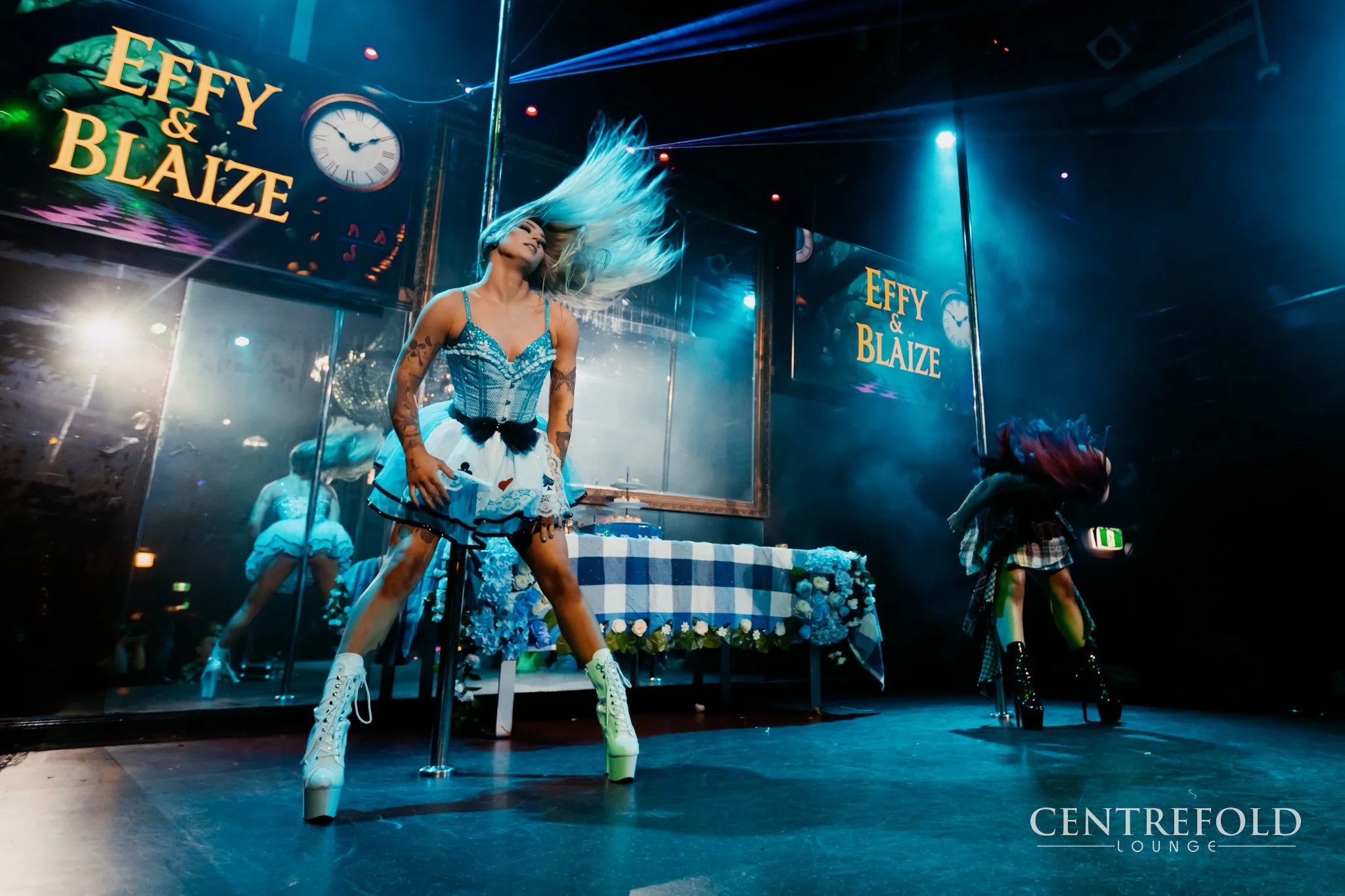 A performer with long blue hair dancing on stage at the Centrefold Lounge, with a backdrop showing the names Effy & Blaize and a clock, wearing a blue and white costume with high white boots, in a nightclub setting with colorful lighting.