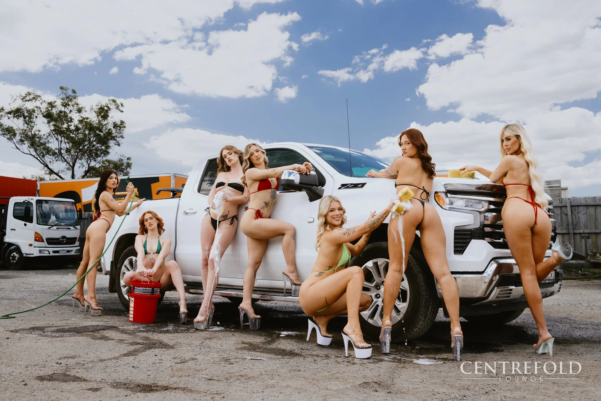 Multiple women in bikinis washing a white pickup truck with sponges and soap, some wearing high heels, outdoors on a gravel lot under a cloudy sky, with a second truck and orange building in the background.
