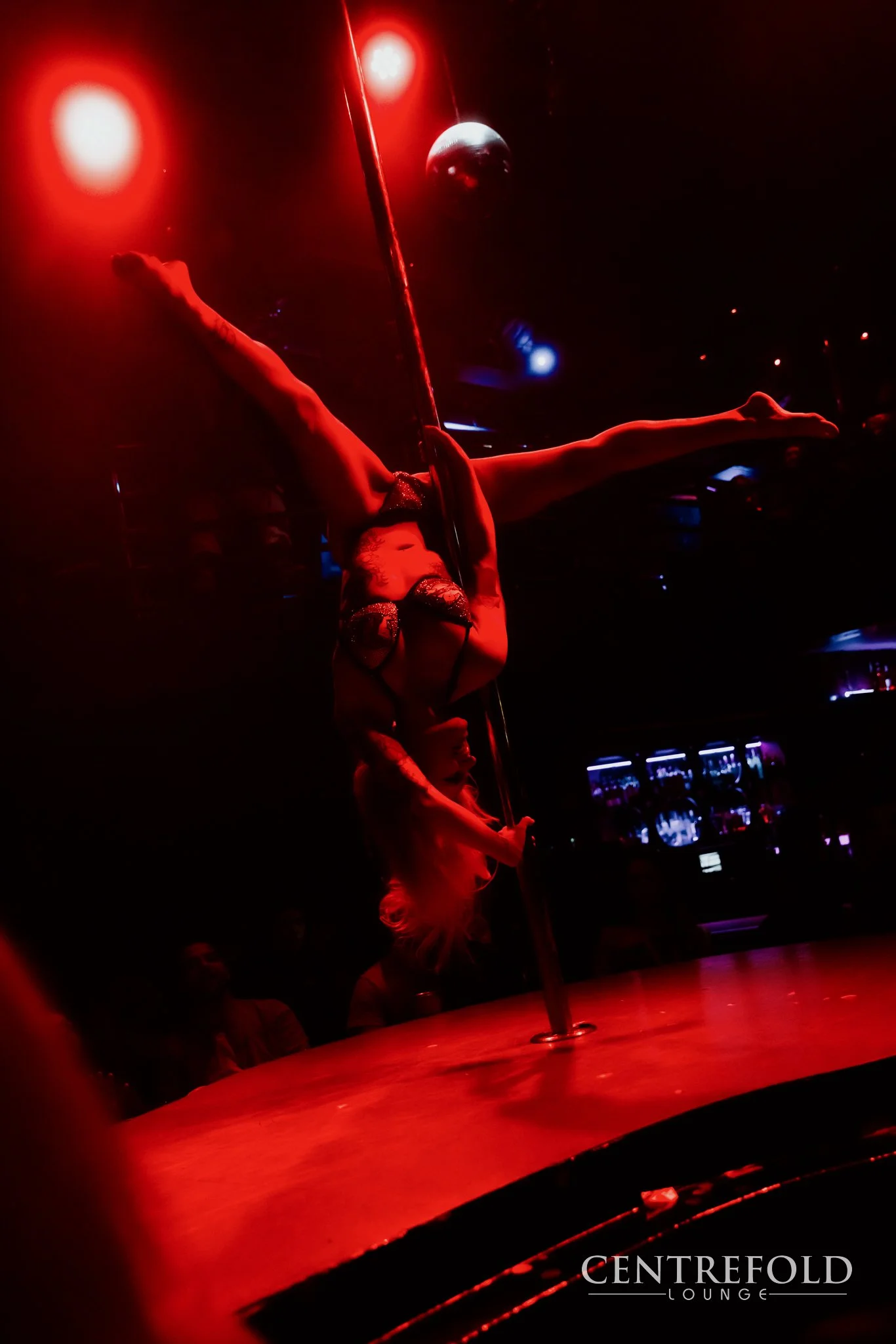 A pole dancer performing in a dimly lit nightclub with red and blue lighting, on a stage with a reflective floor, with the logo 'Centrefold Lounge' visible in the bottom right corner.