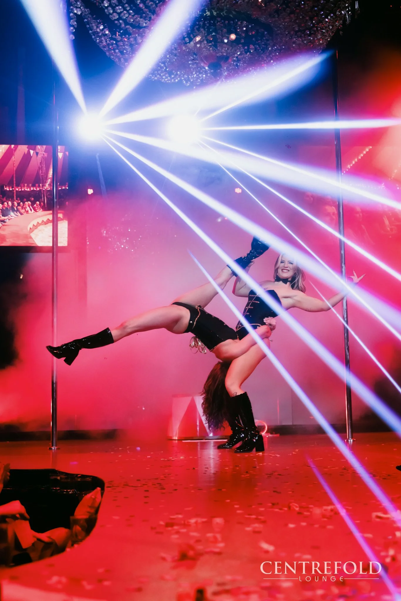 Two women performing a pole dance on a stage with colorful spotlights and confetti, in a lounge setting named CENTREFOLD LOUNGE.