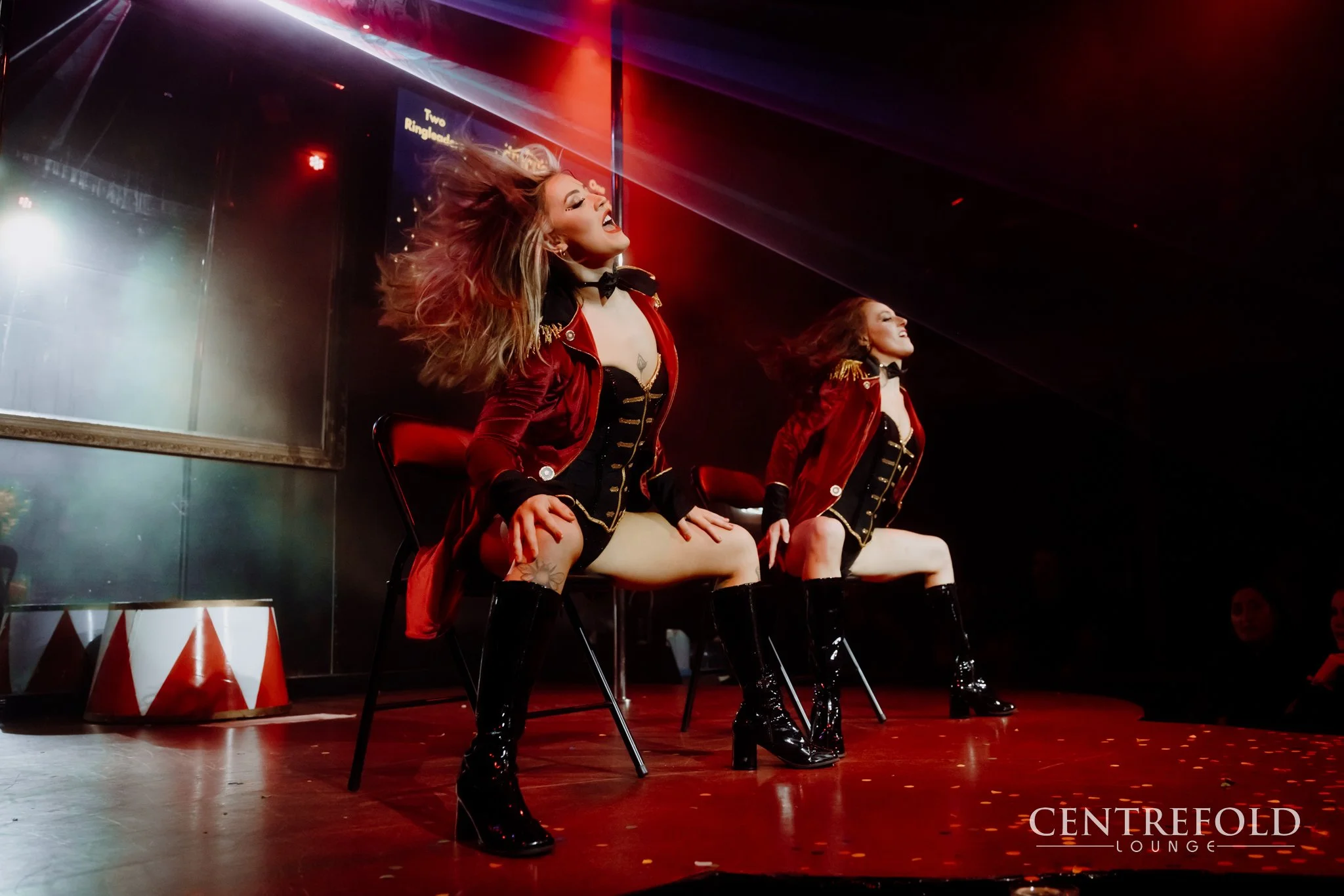 Two women performing on stage at Centrefold Lounge, dressed in red velvet jackets, black corsets, and high black boots, with hair flowing and expressive movements.