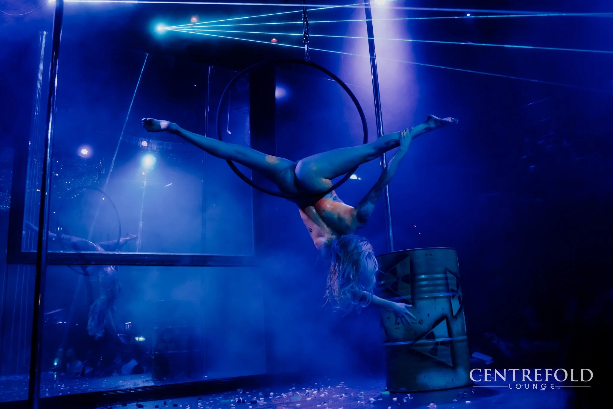 A female performer in a blue costume is hanging upside down from a circular aerial hoop with her legs spread apart and one arm reaching down, on a stage with blue lighting and fog effects, in front of a large mirror at Centrefold Lounge.