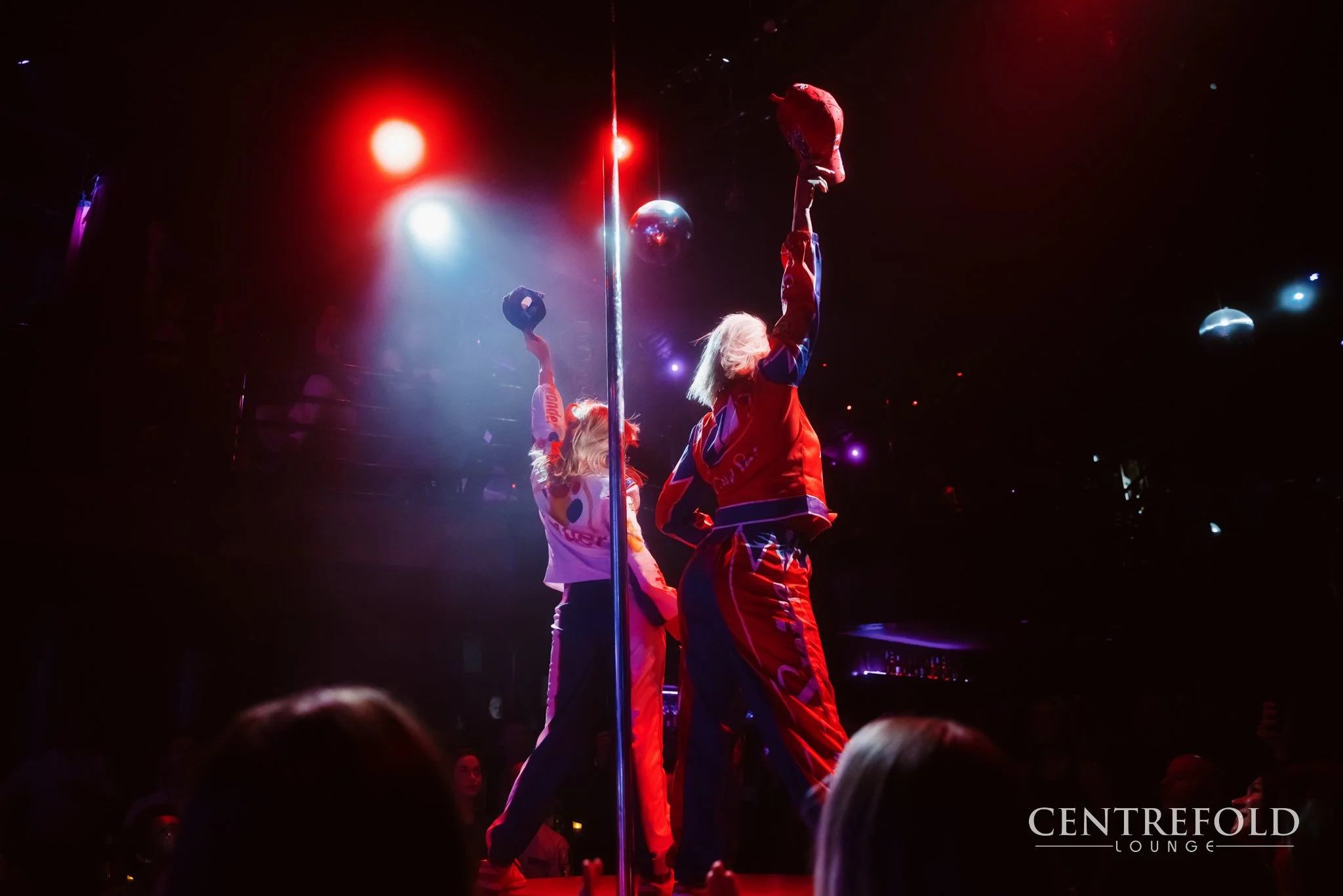 Two performers with blonde hair on a dimly lit stage, holding hats above their heads, near a pole, with colorful lights and disco balls overhead at CENTREFOLD LOUNGE.