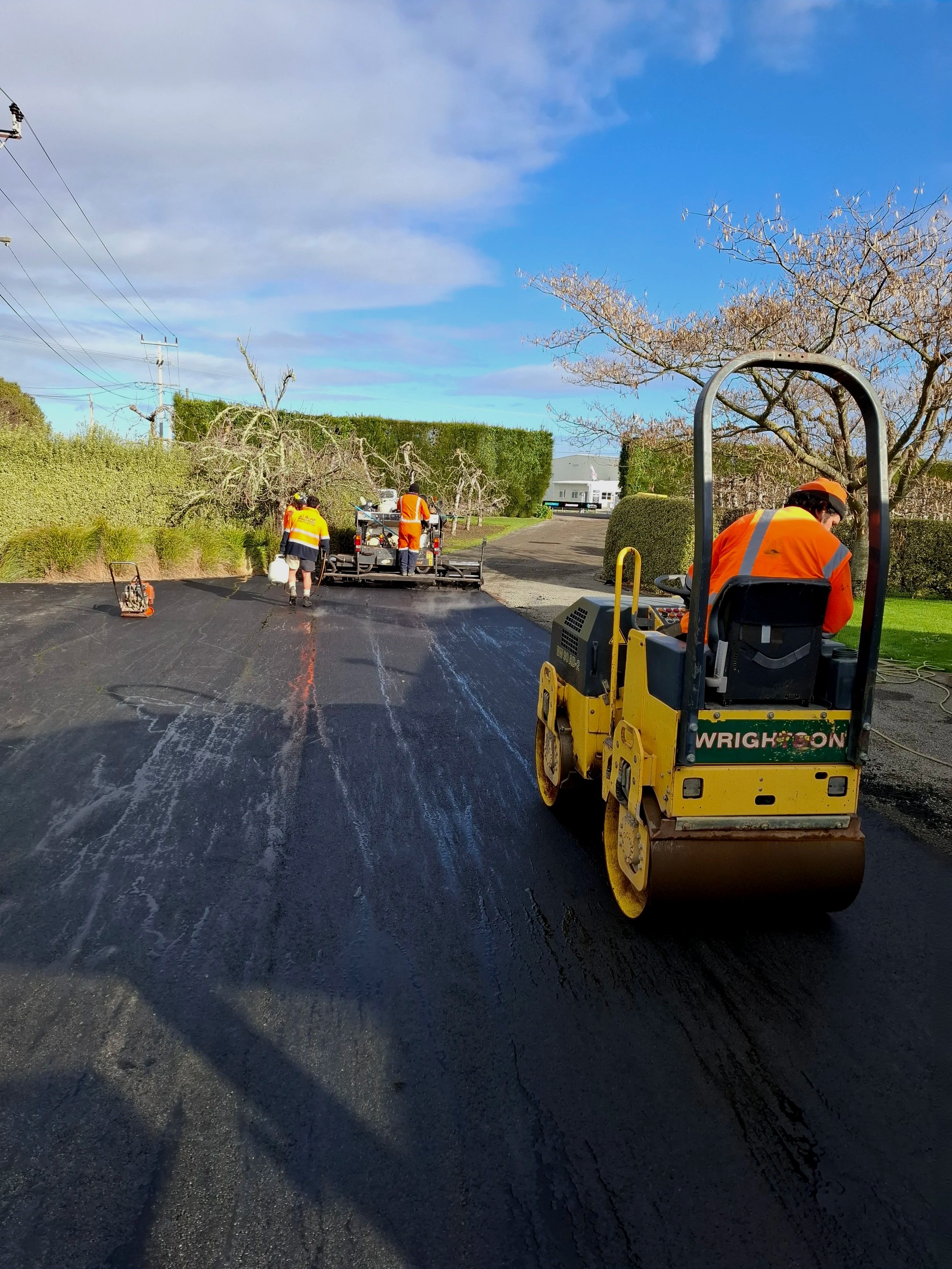 Wrightson Contracting - Napier, Hastings and wider Hawke's Bay. Workers in orange safety vests operating road paving equipment on a newly paved asphalt road. There are trees and hedges along the sides of the road and a partly cloudy sky above.