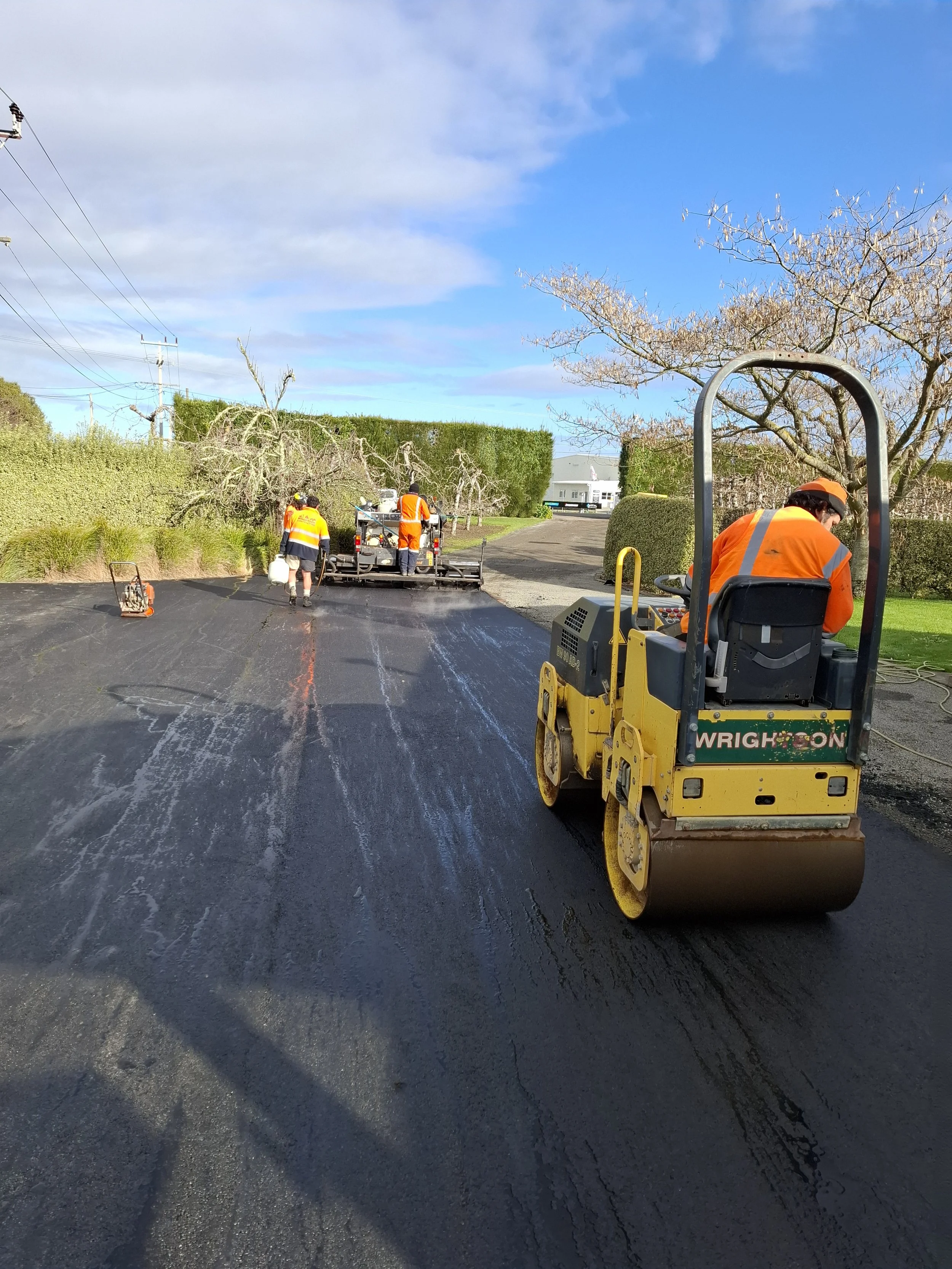Workers in orange uniforms paving a road with a yellow vibratory roller and asphalt paving machine, with trees and a building in the background.