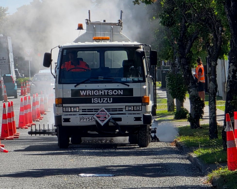 Wrightson Contracting - Napier, Hastings and wider Hawke's Bay. City street construction scene with a white road maintenance truck, orange cones, workers in safety vests, and trees along the sidewalk.