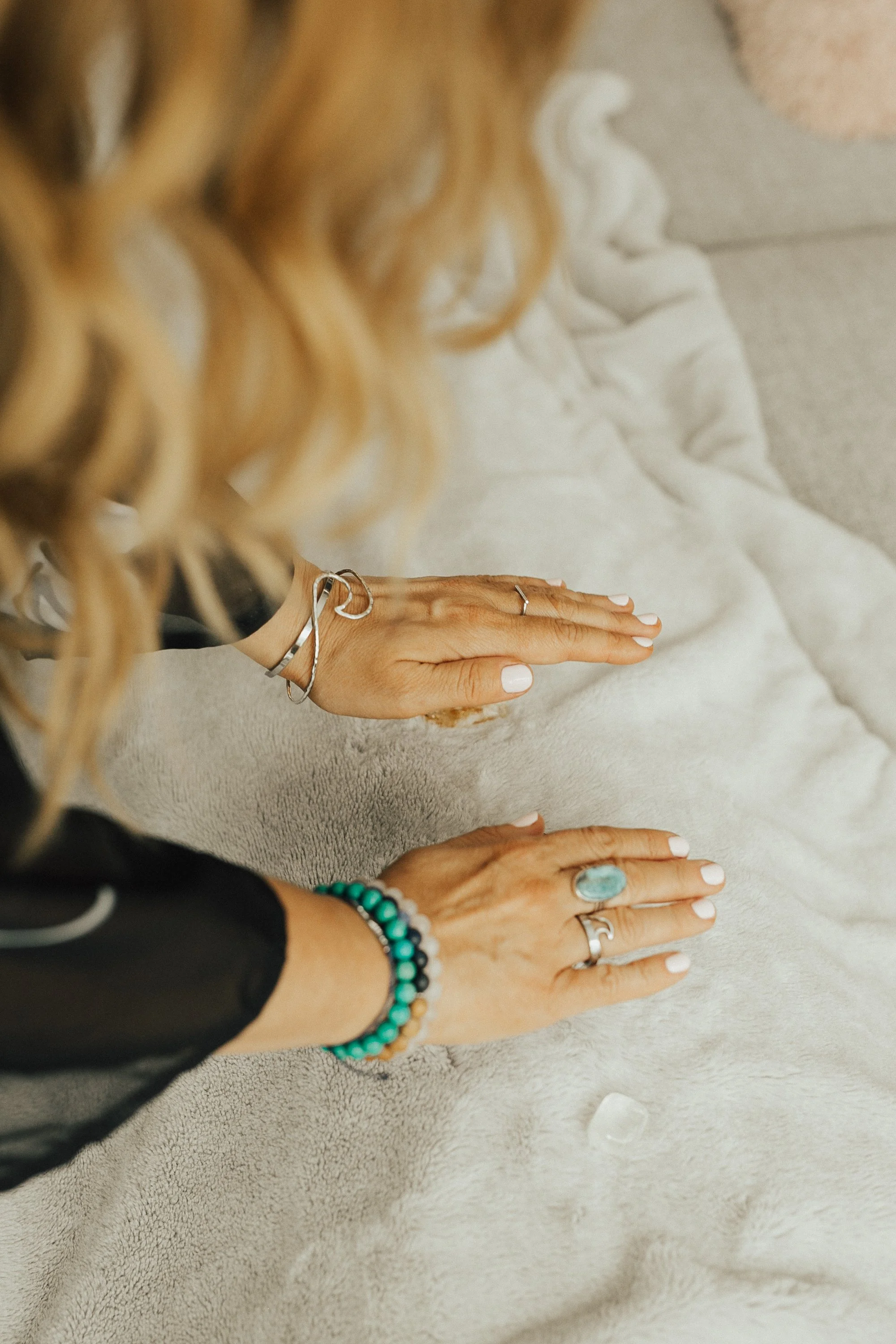 Person with blonde curly hair places hand on a soft beige blanket, wearing various jewelry including rings and bracelets, with part of white clothing visible.