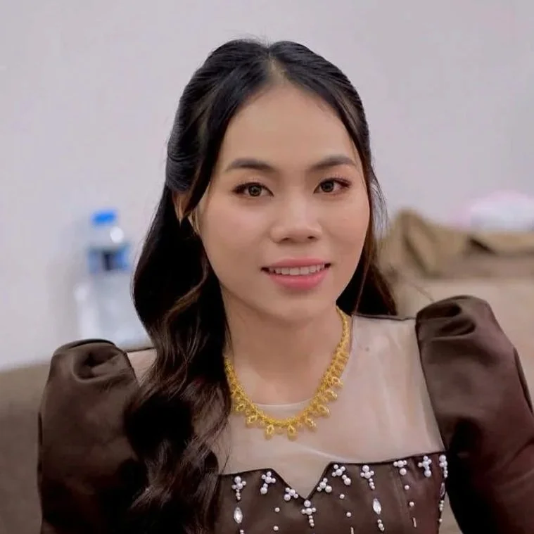 A young woman with long black hair, wearing a light blue top with a white collar, smiling softly at the camera against a light-colored background.