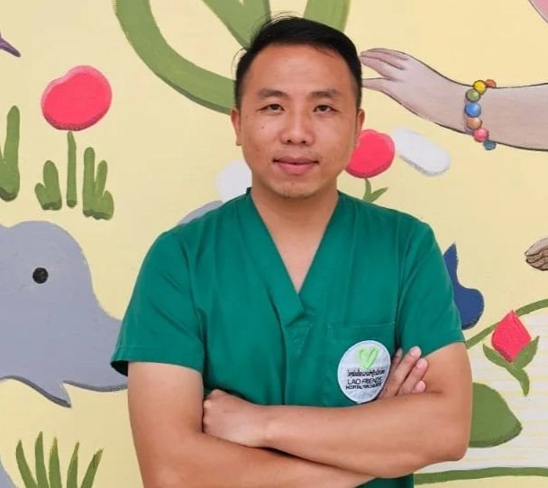 A young male healthcare professional in green scrubs, smiling, wearing a red stethoscope around his neck, indoors with natural light.
