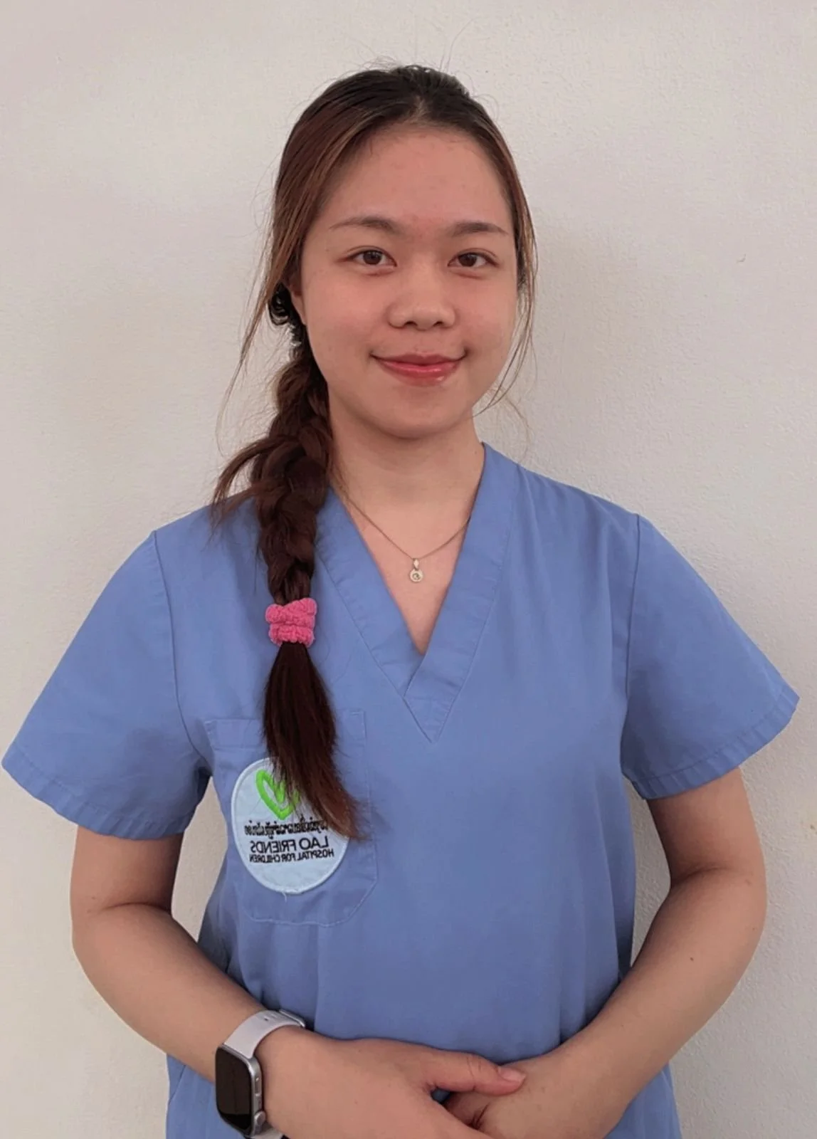 A young woman with long black hair braided over her right shoulder, wearing a white T-shirt with a green heart logo, standing in front of a yellow wall.