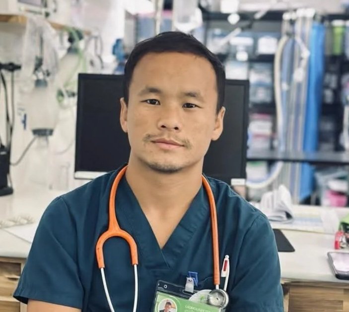 A young male healthcare professional wearing a green scrub top and stethoscope, smiling outdoors.