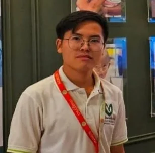A young man with black hair wearing a black suit, white shirt, and black tie against a solid blue background.