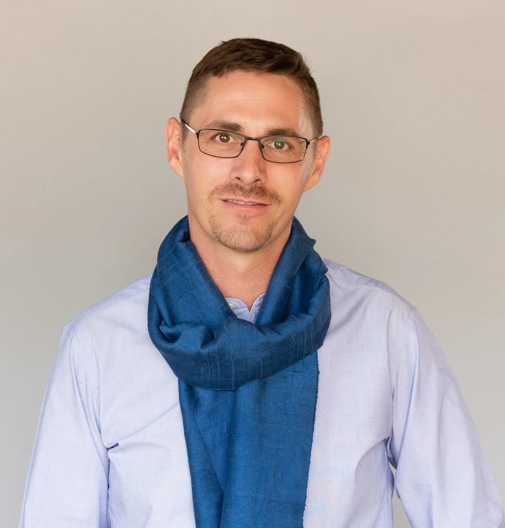 A man with glasses and short brown hair wearing a light blue dress shirt and a blue scarf poses against a plain light background.