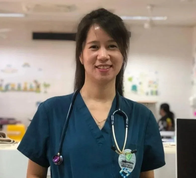 A female healthcare worker in blue scrubs with a stethoscope around her neck, standing indoors against a beige wall with framed artwork.