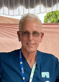 A smiling elderly man with gray hair and glasses wearing a navy blue medical uniform with a hospital badge and a phone in the pocket, standing against a white wall.