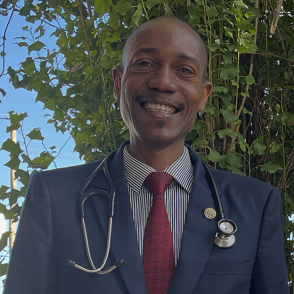 A smiling elderly man with gray hair and glasses wearing a navy blue medical uniform with a hospital badge and a phone in the pocket, standing against a white wall.