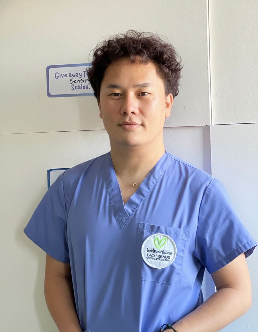 A person wearing light blue medical scrubs with an ID badge, standing indoors against a wooden wall.