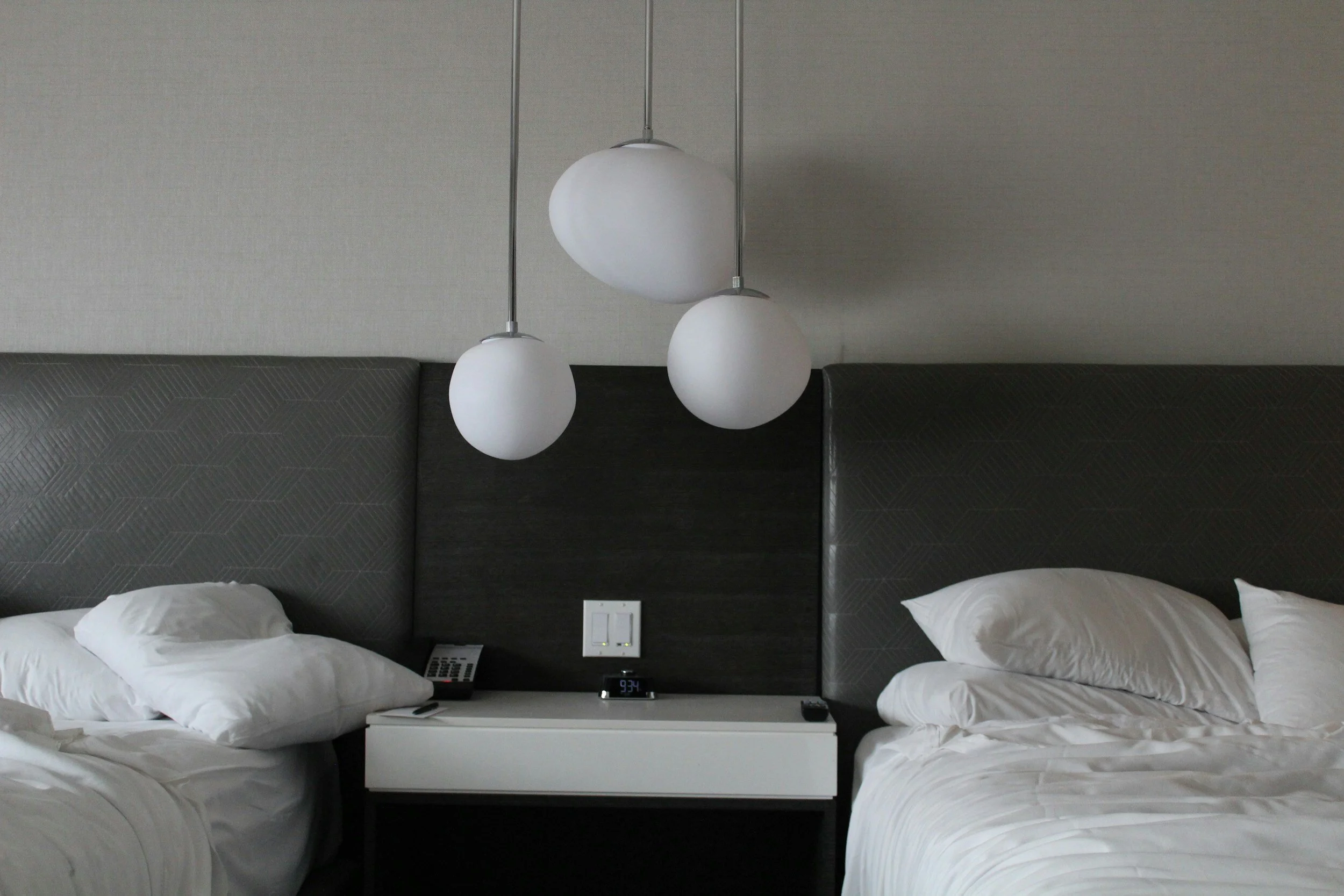 Two unmade hotel beds with white linens and pillows, a black headboard, a white nightstand with a phone, clock displaying 3:34, and a remote, with three modern white pendant lights hanging above.