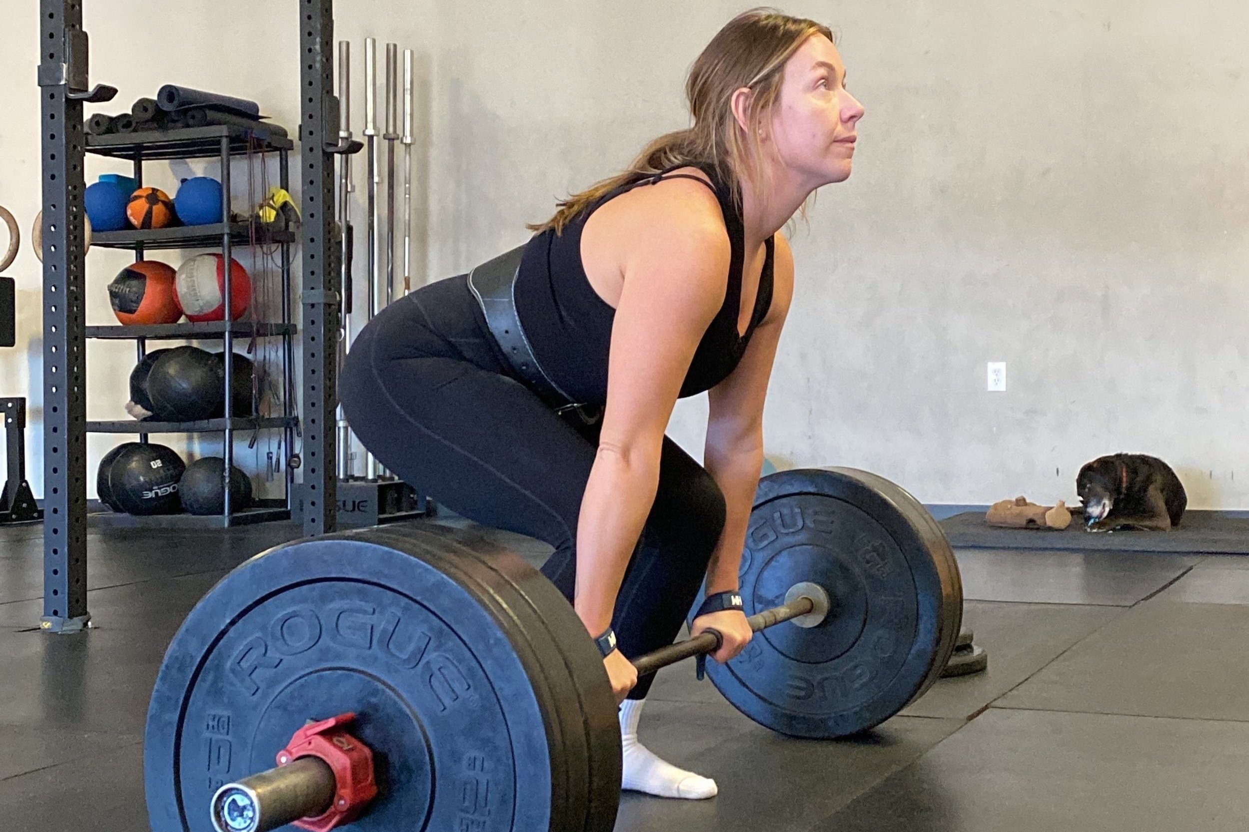 Woman lifting a barbell in a gym, with a dog lying on a mat in the background.