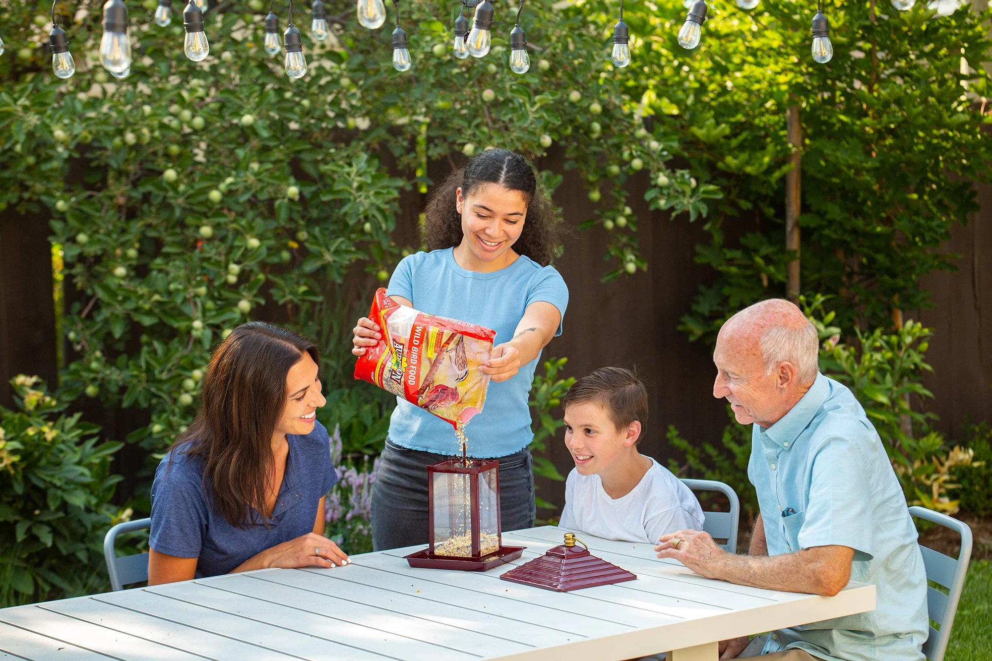 Grandfather, mother, teenager girl and young boy sitting around backyard table watching teenage girl fill bird feeder with bird seed.