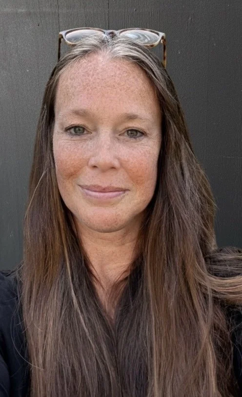 Smiling woman with long hair indoors, next to a window, with blinds and greenery decoration on the wall.