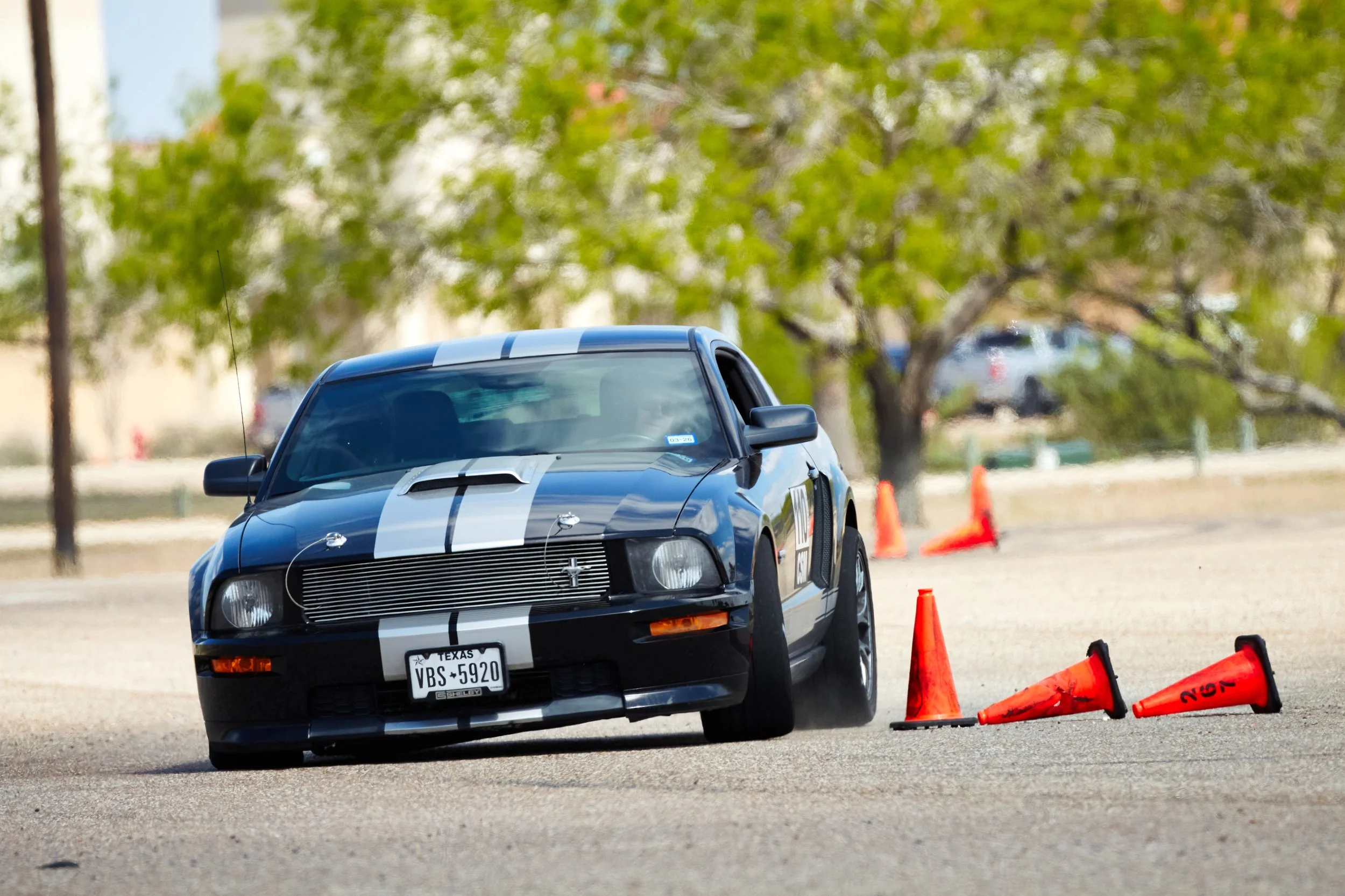 A black race car with white racing stripes on a track, navigating around orange traffic cones, with a background of trees and parked cars.