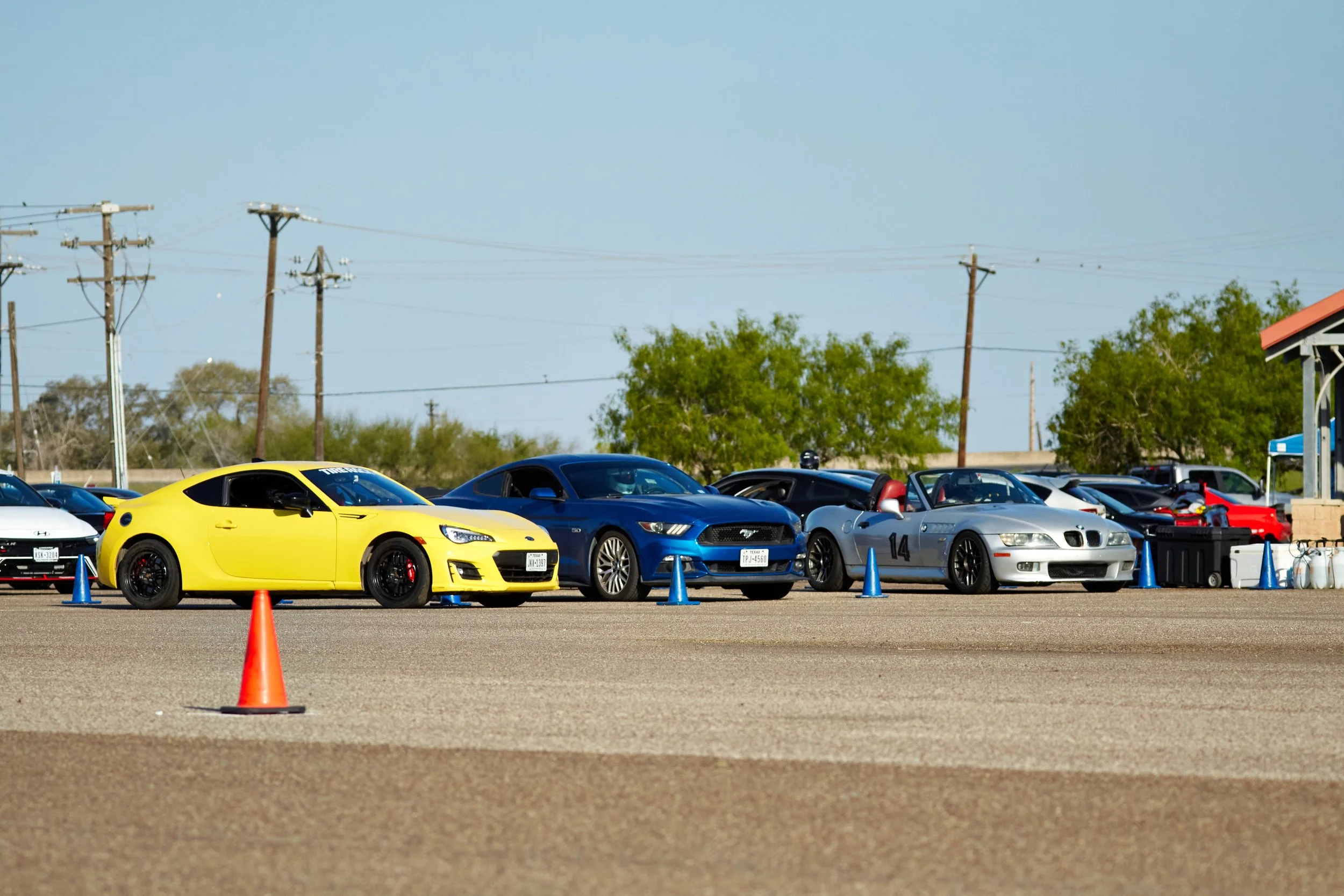A row of sports cars parked behind blue cones on a paved lot, with a yellow car on the left, a blue car in the middle, and a silver car with a race number on the right, under a clear sky with power lines and trees in the background.