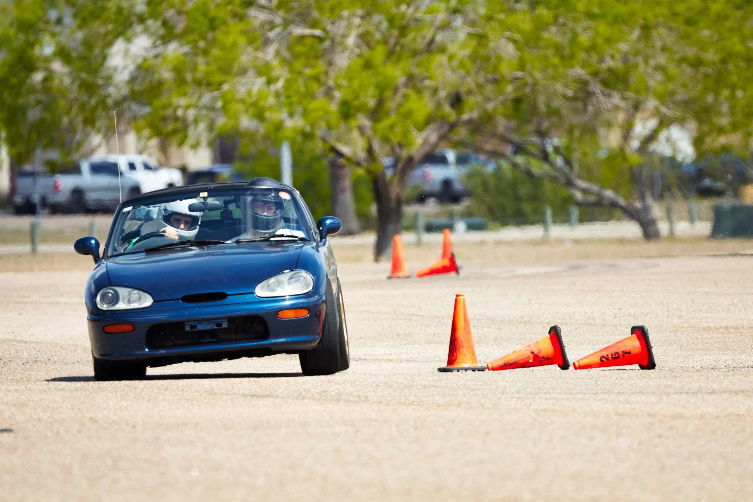 Blue convertible car driving around orange traffic cones on a track with trees in the background.