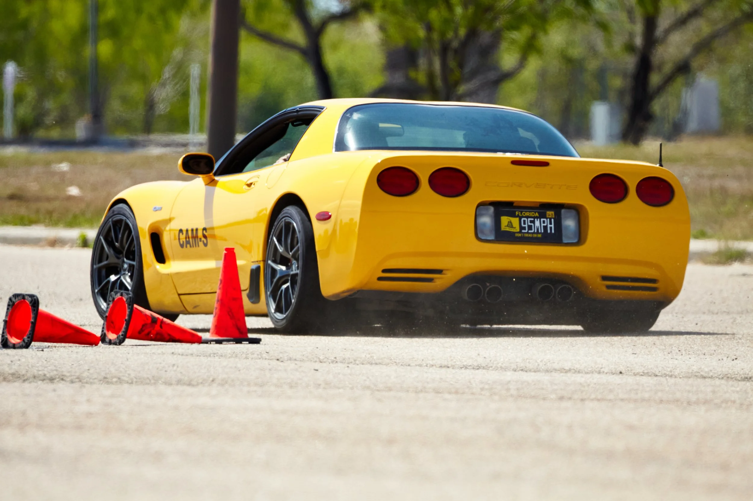 A yellow Chevrolet Corvette sports car with Florida license plate reading '95 MPH,' parked on an empty lot, surrounded by orange traffic cones and safety signs, with trees in the background.