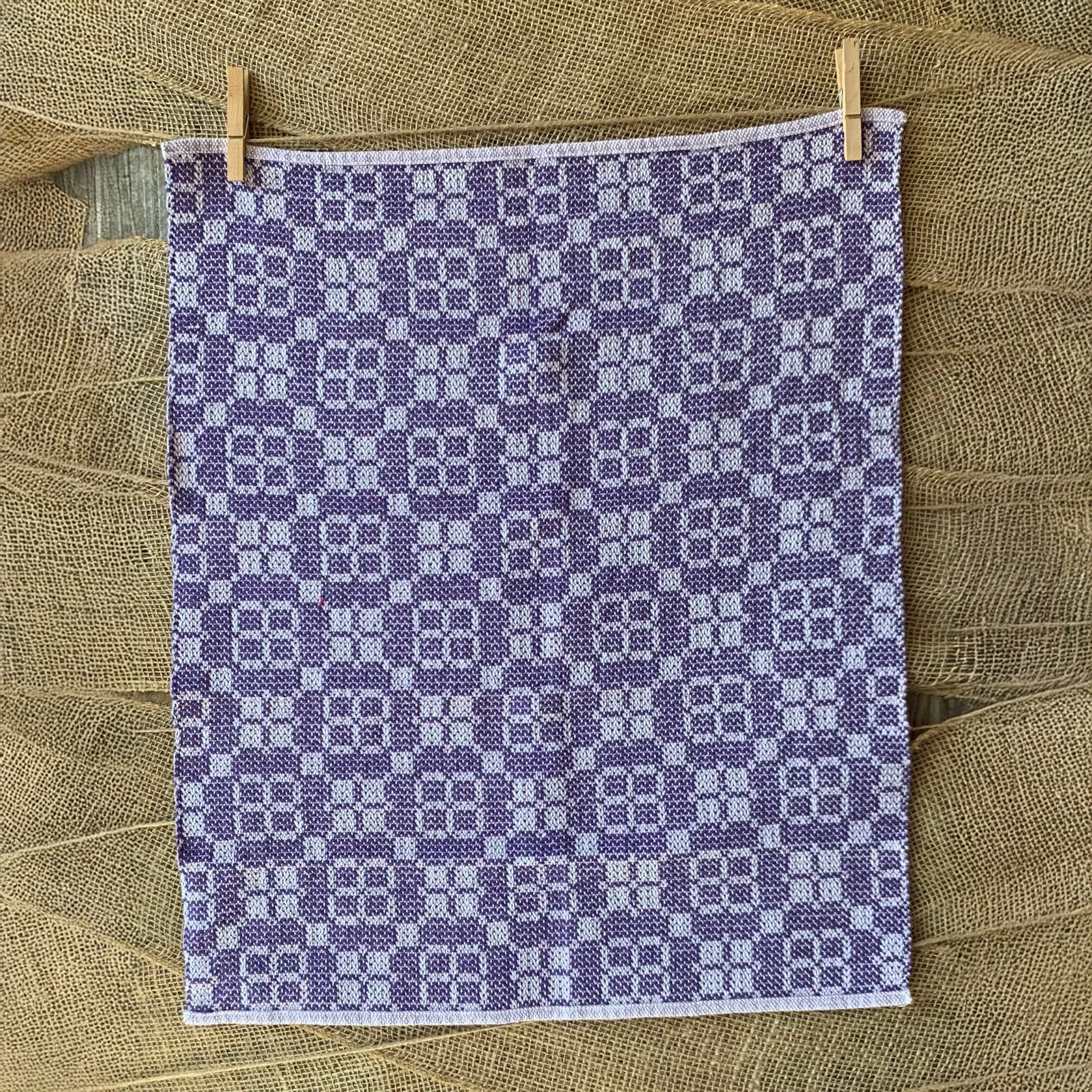 A purple and white dishtowel, hanging on a textured surface with clothespins.