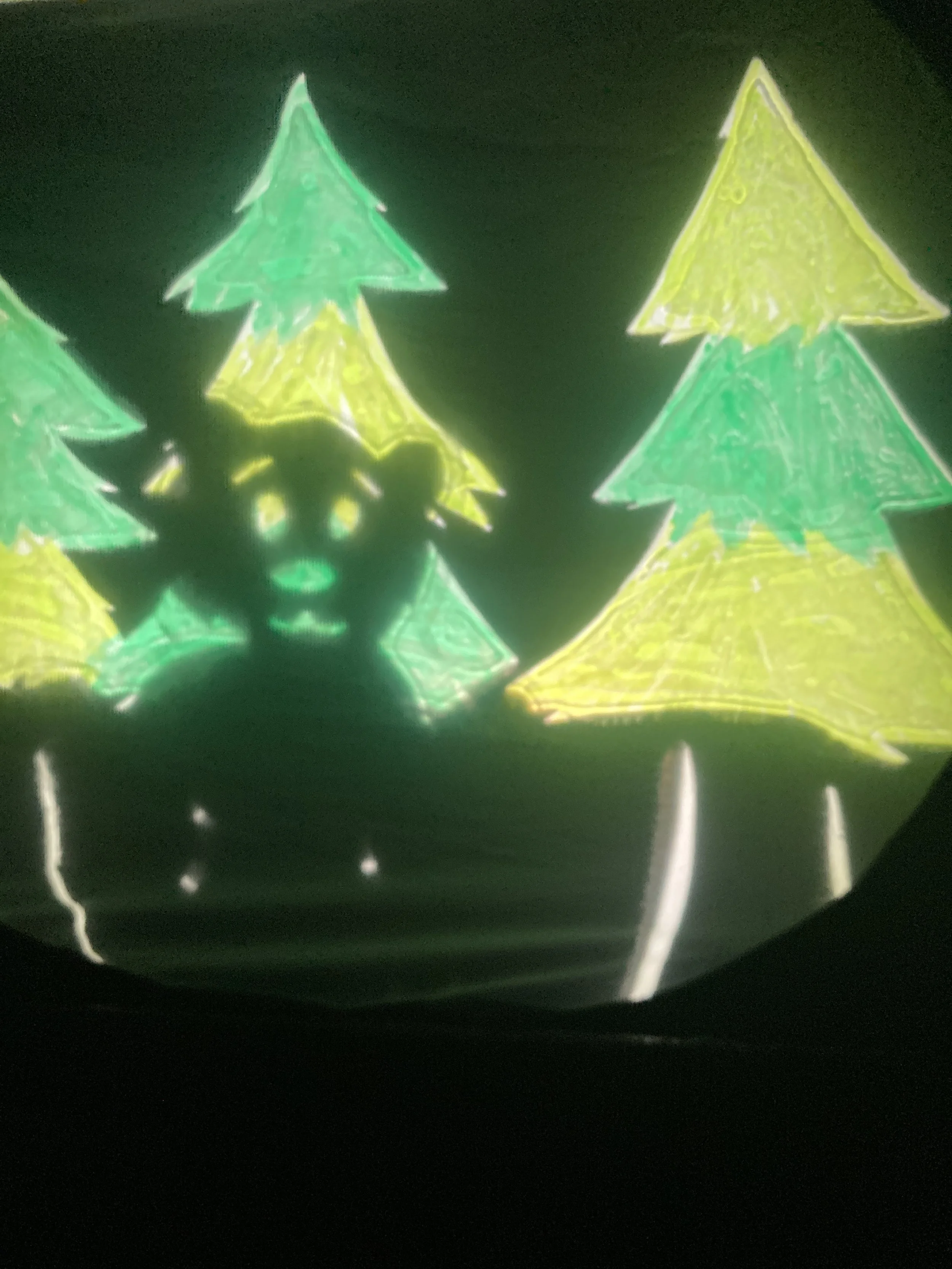 Alt Text: A shadow puppet teddy bear peaks through the green branches of a forrest