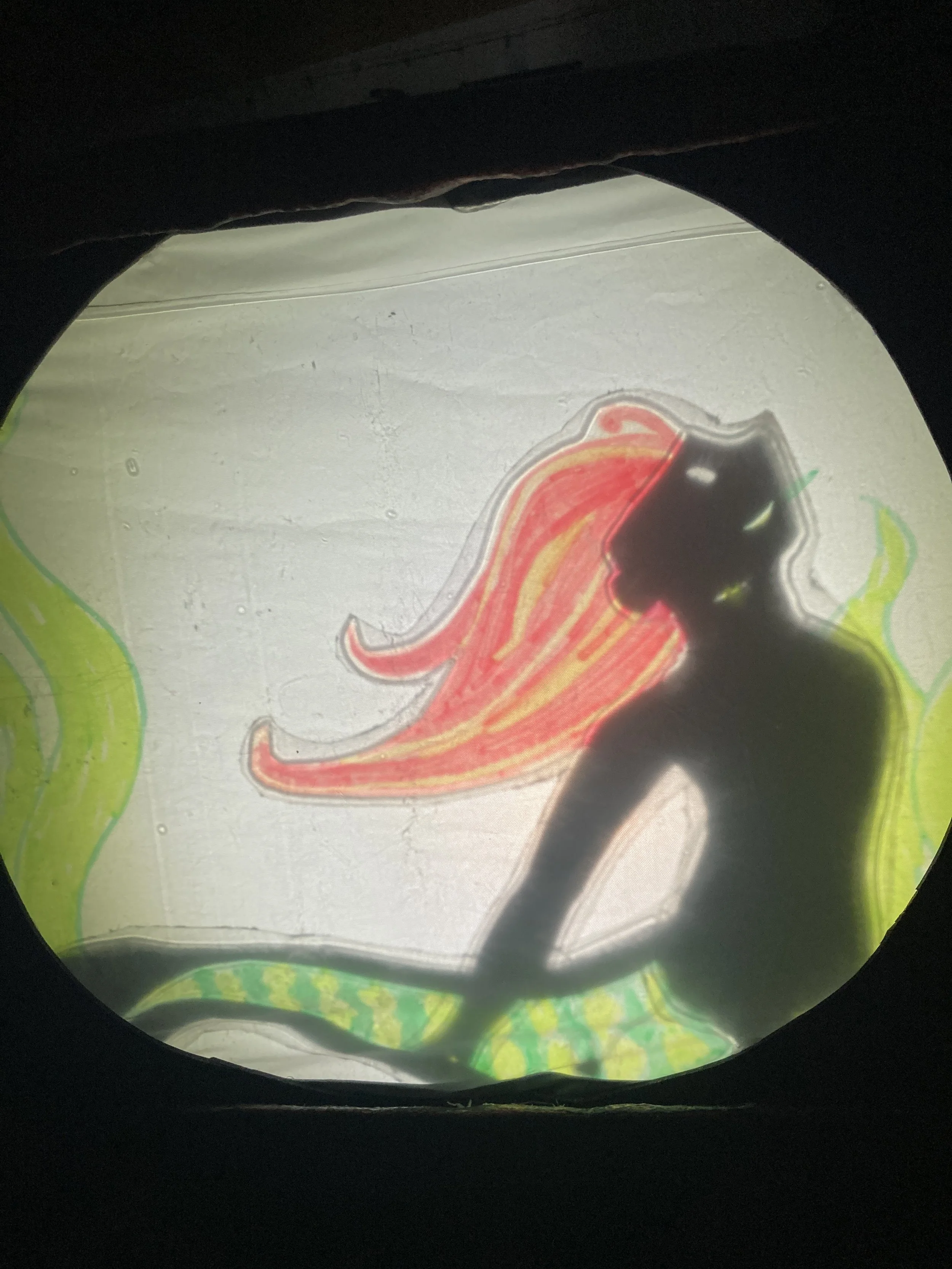 Alt Text:A shadow puppet mermaid with red hair and a green fish tail looks up