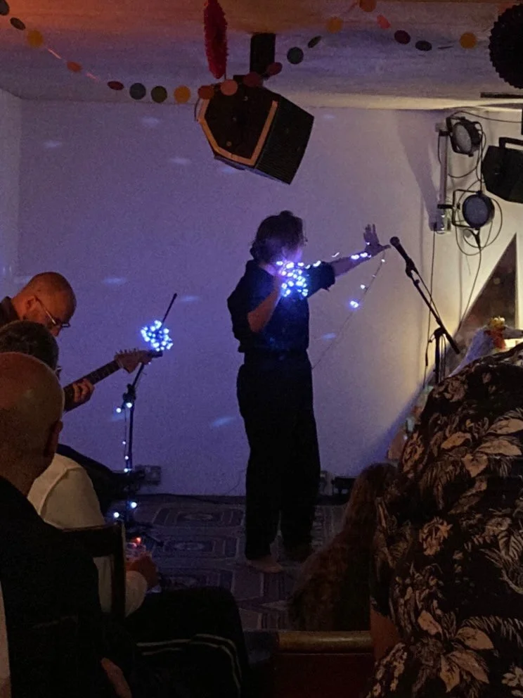 A photo from Scratch|Meet 4. Joni is standing with a bundle of LED lights in her right hand. Her left arm is out stretched behind her with a string of LED. Behind her is a mic stand which is also wrapped up in LEDs. The LED's have a white-ish blue-is