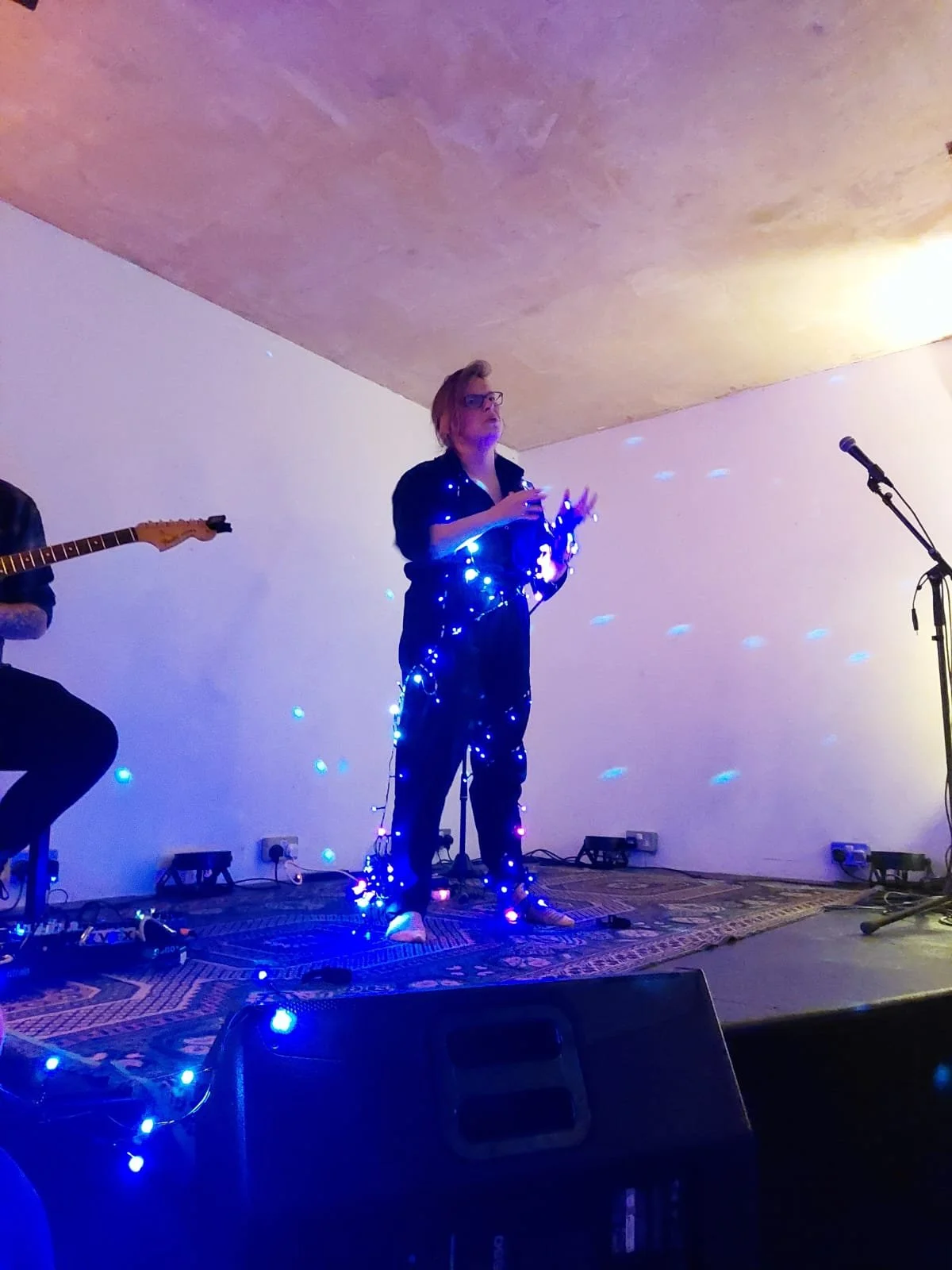 A photo from Scratch|Meet 4. Joni is standing wrapped up in LED's in the middle of a monologue. The LED's have a white-ish, blue-ish glow and look like a constellation of stars 