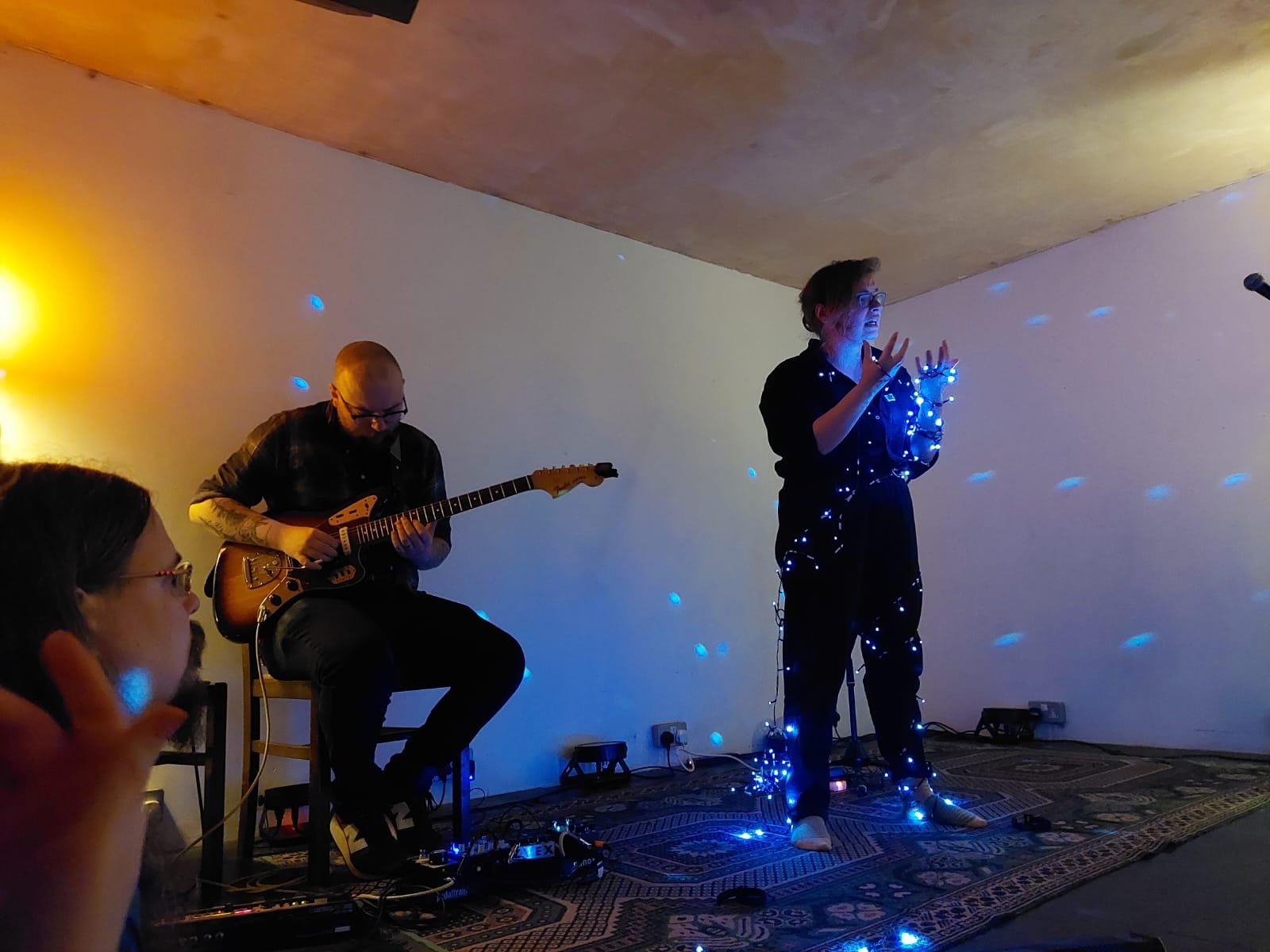 Photo from Scratch|Meet 4. Joni is standing wrapped up in LED lights. They have a white-ish blue-ish glow. Iain Scott is playing a guitar in the corner. 