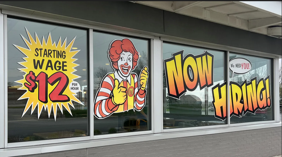 A fast-food restaurant window displays large signs reading “Starting Wage $12 per hour” and “Now Hiring,” alongside a picture of Ronald McDonald giving a thumbs-up.