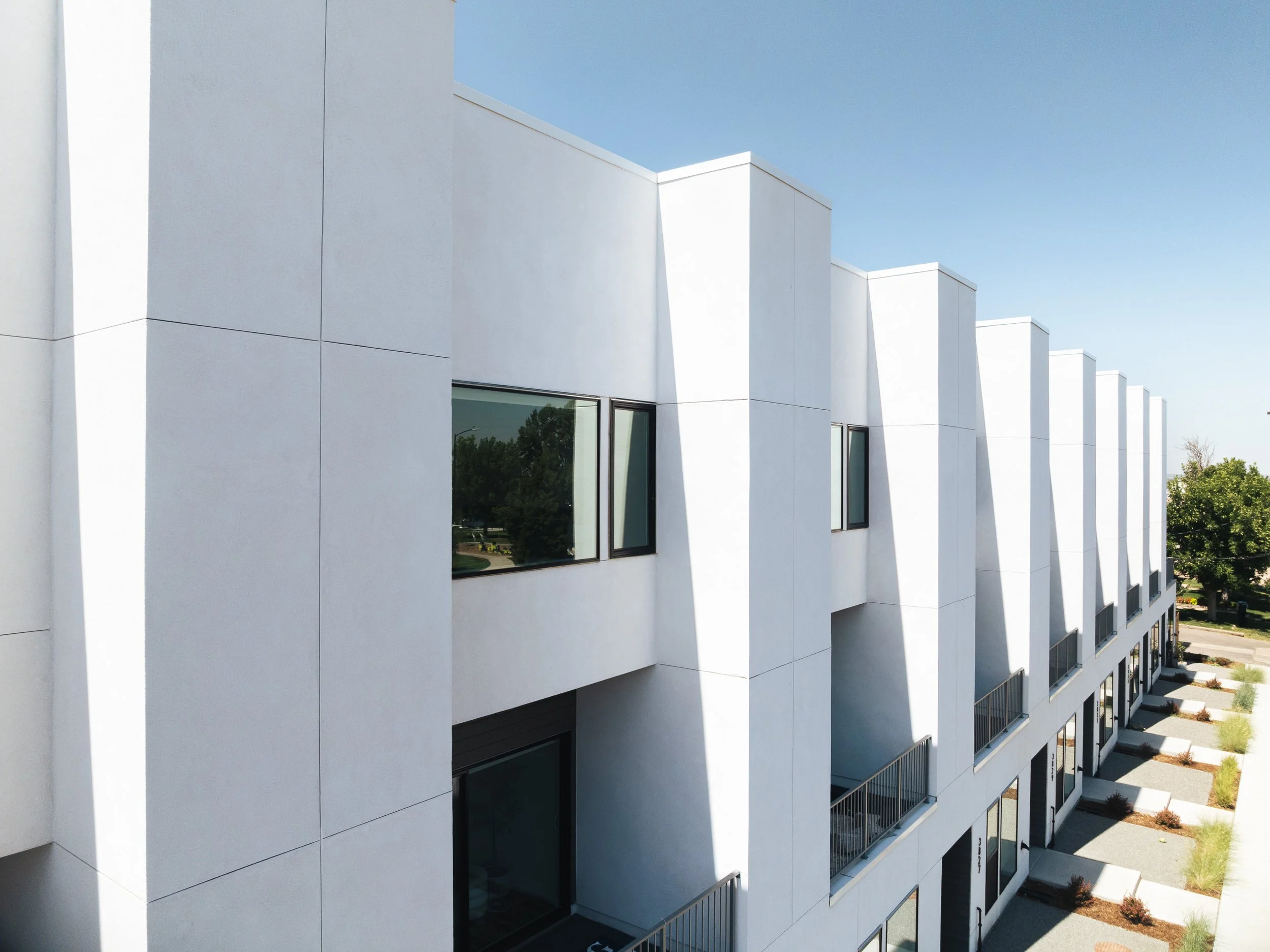 Modern white residential building with multiple stories, large windows, and small balconies, set against a clear blue sky.