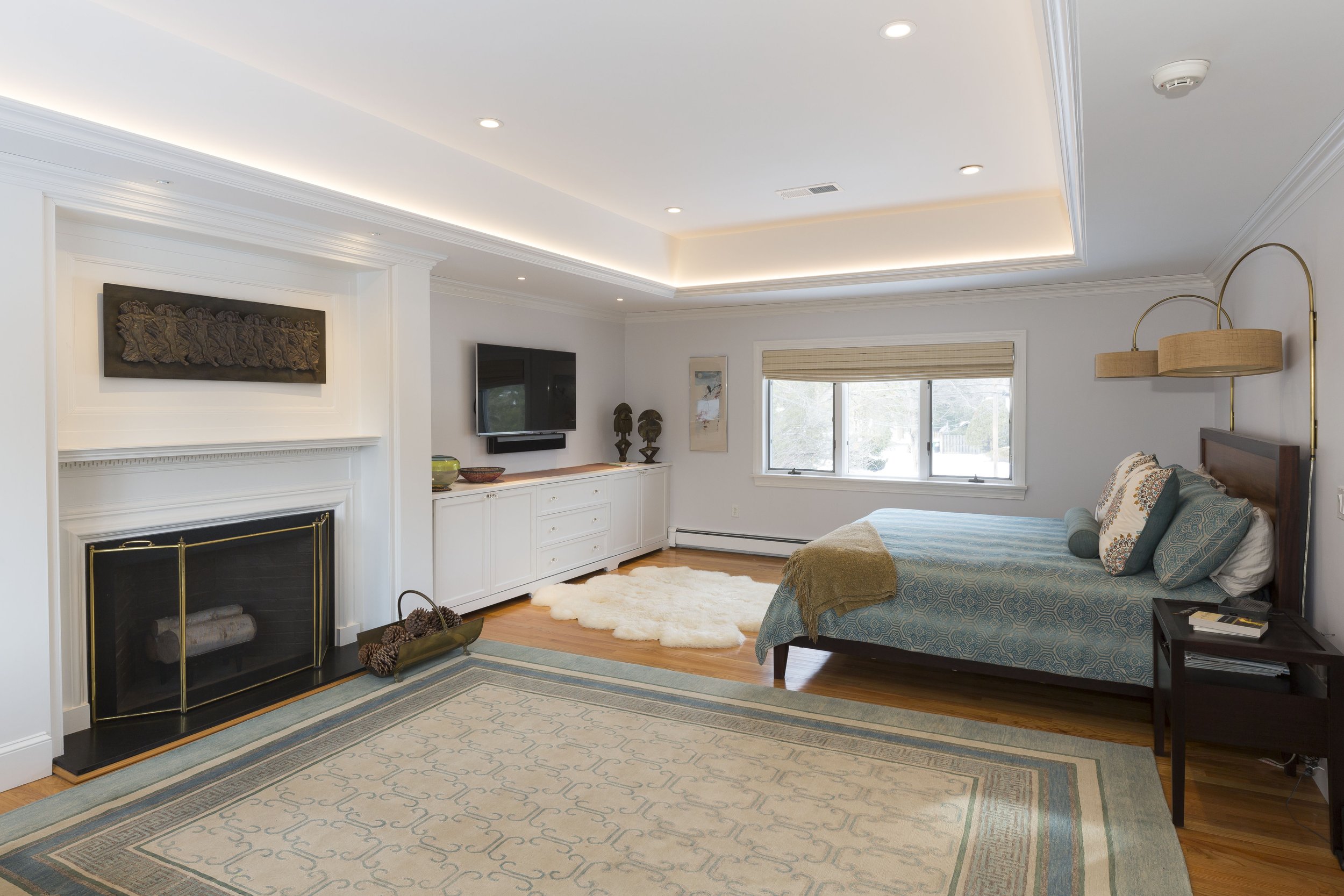A modern living room with a white ceiling, crown molding, recessed lighting, and a large window. There is a fireplace with a decorative sculpture above it, a wall-mounted television, and a low white cabinet. The room has a bed with patterned pillows, a beige blanket, and a fluffy white rug on a hardwood floor. A colorful rug is partially visible in the foreground.