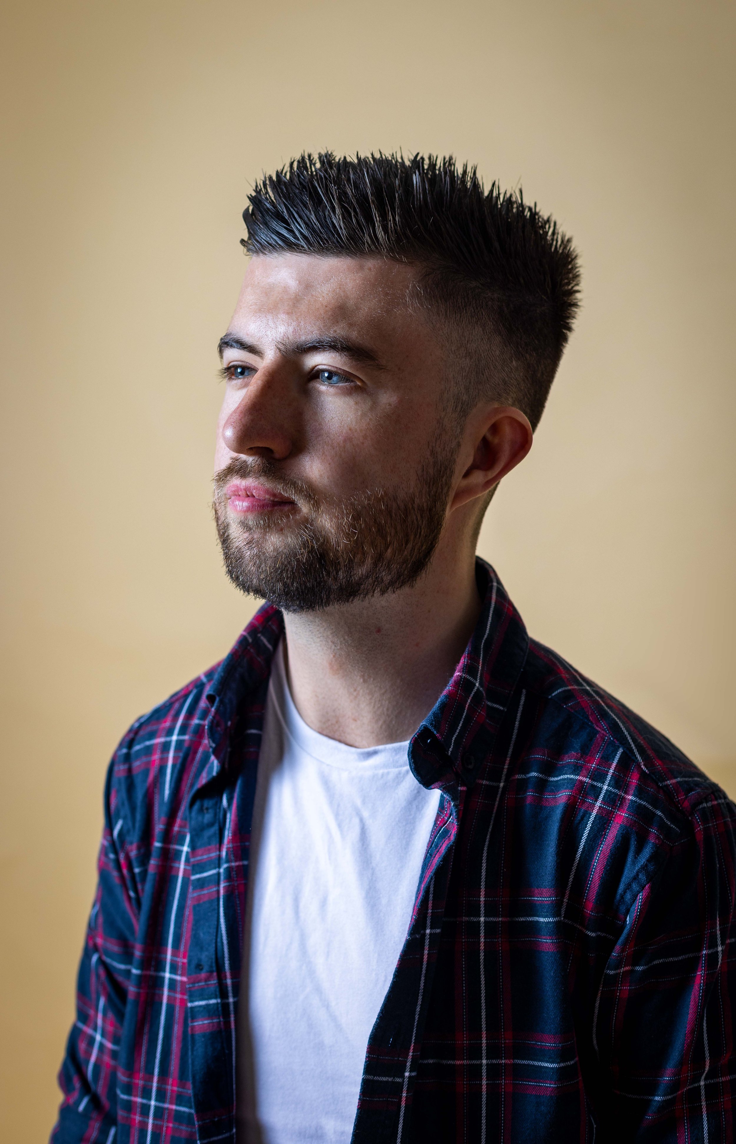 Man with short beard and spiked hair wearing a plaid shirt and white t-shirt against a plain background.