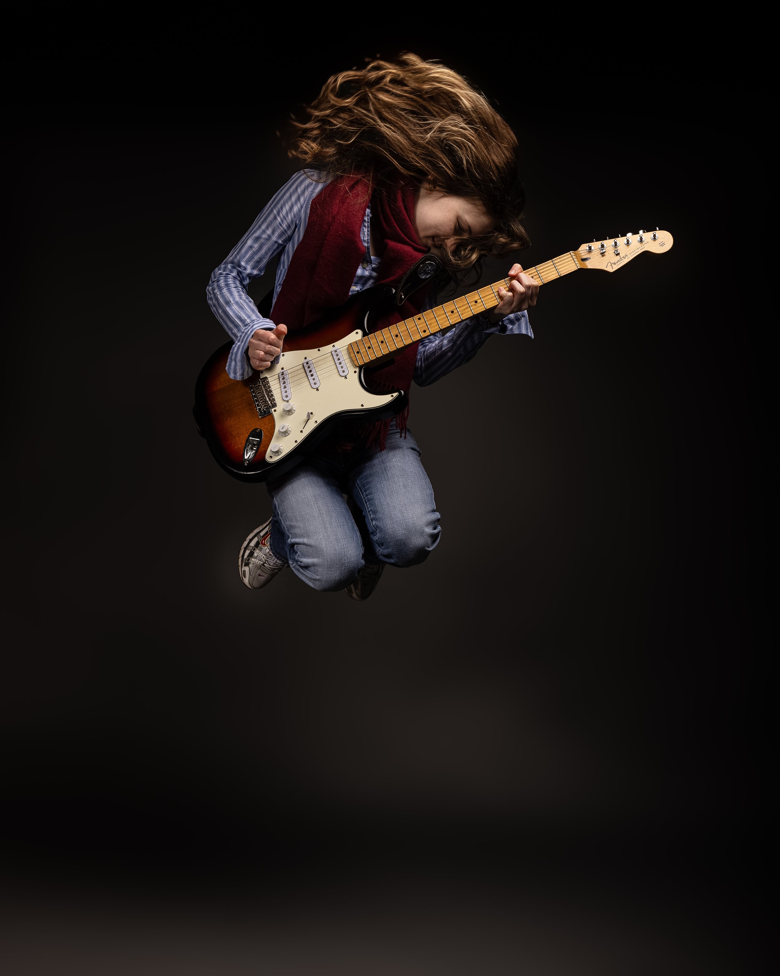 Person with long hair jumping playing electric guitar, wearing jeans and a scarf, black background