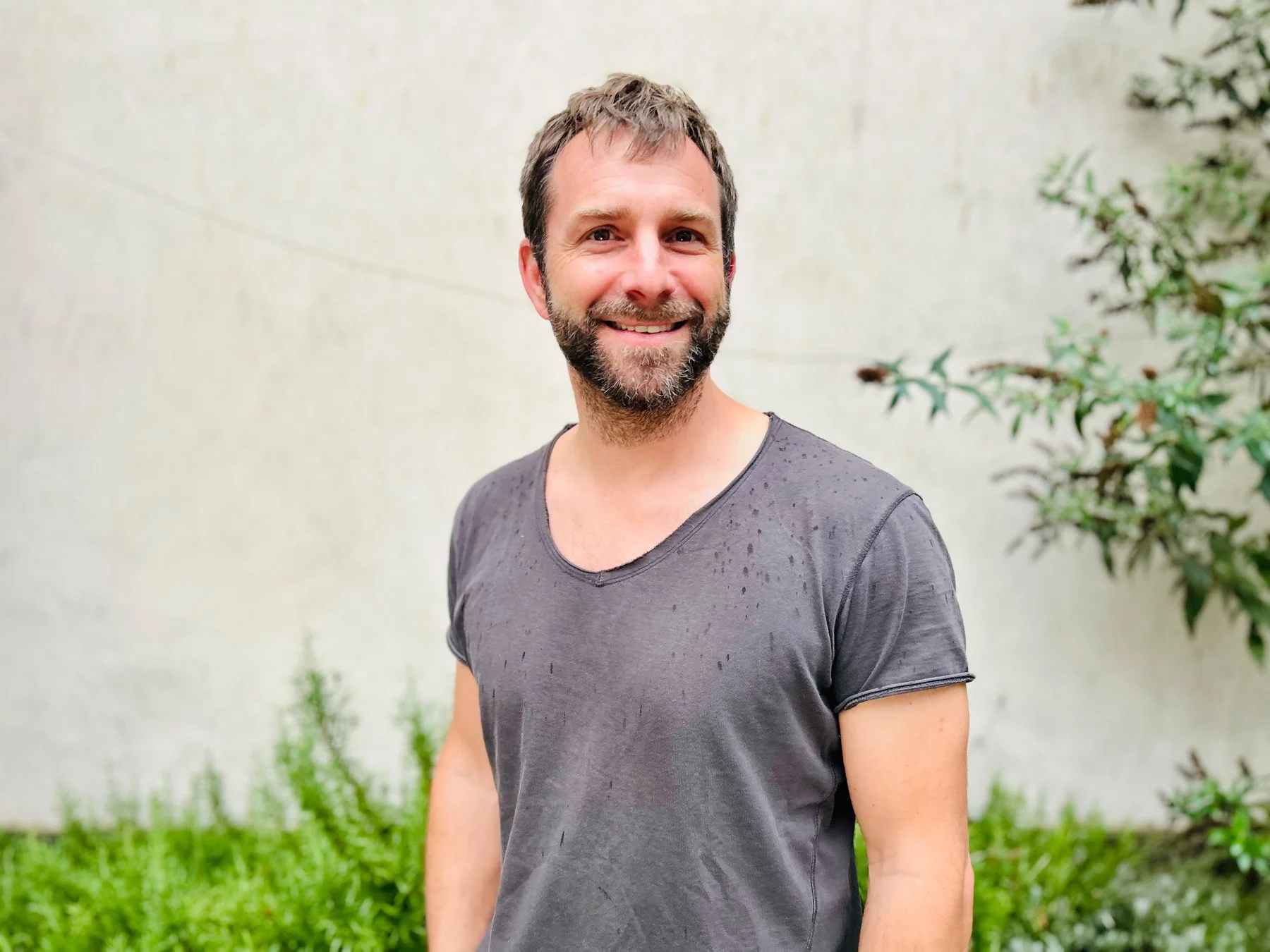 Lächelnder Mann mit kurzen braunen Haaren und Bart, trägt ein nasses graues T-Shirt, steht vor einer weißen Wand und grünen Pflanzen draußen.