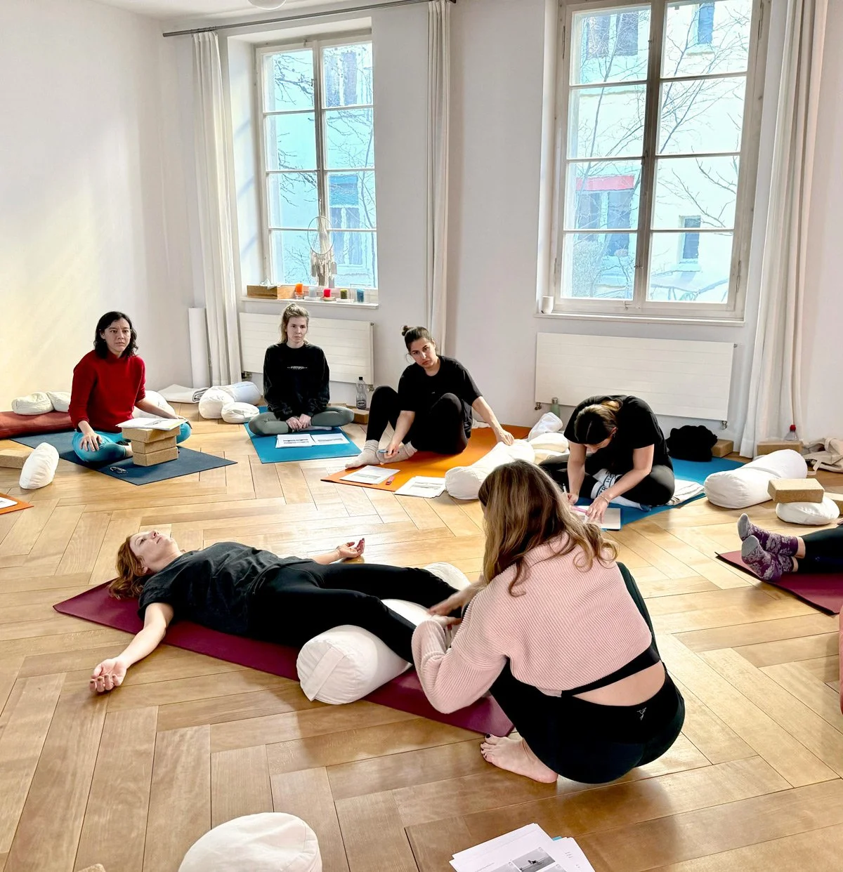 Eine Gruppe von Frauen sitzt auf Yogamatten im Raum, eine Frau liegt auf einer Yogamatte in der Mitte, während eine andere sie berührt. Der Raum hat große Fenster und Holzboden. Es sieht aus wie eine Yogastunde oder Meditationsklasse.