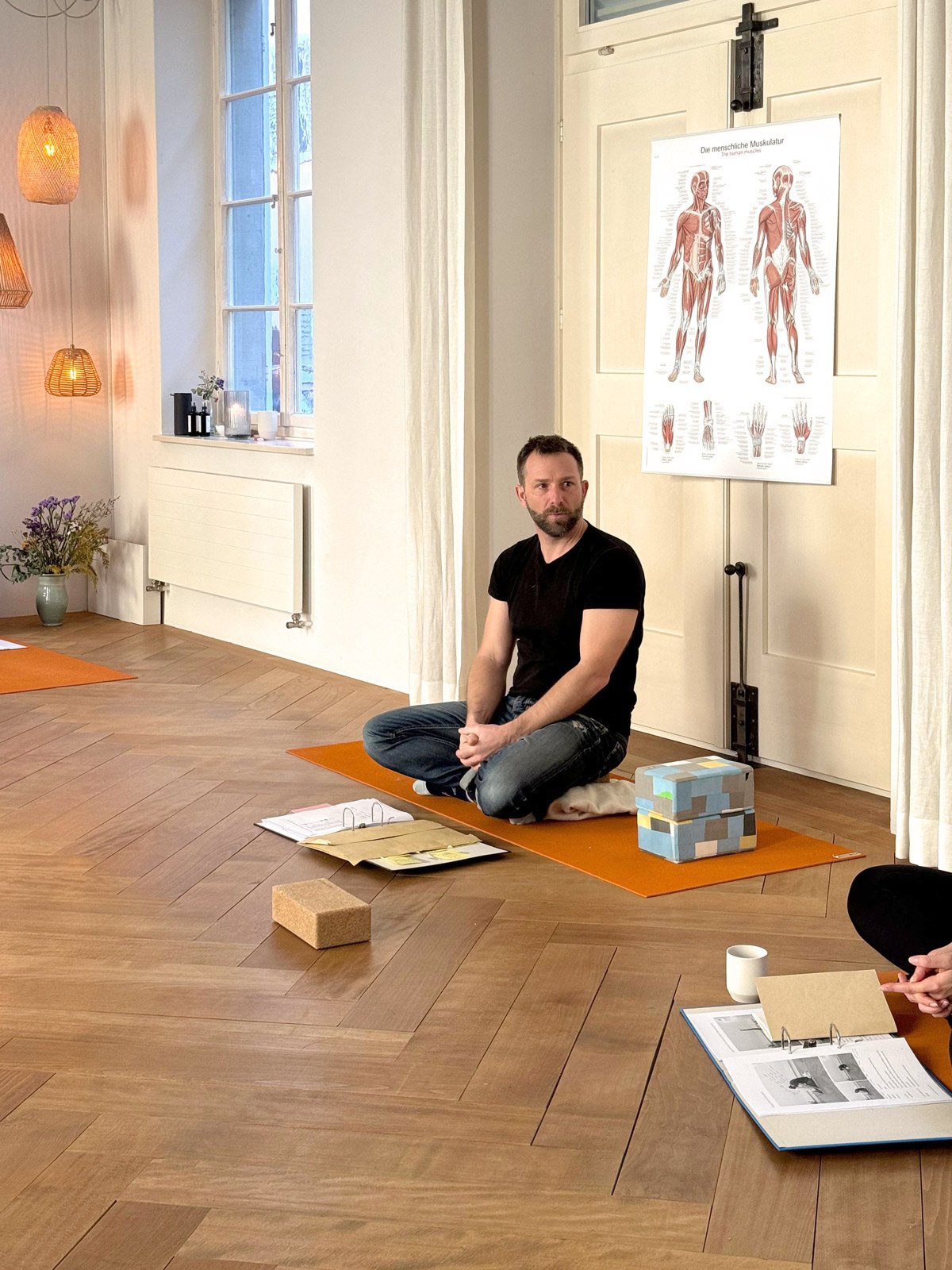 Ein Mann sitzt auf einem Yogamatte bei einem Vortrag über menschliche Muskulatur. Es gibt offene Ordner, Bücher, einen Ziegelstein, Papiere und eine Stempelbox um ihn herum. Im Hintergrund eine weiße Wand mit einem Fenster und einem Poster über die menschliche Muskulatur.
