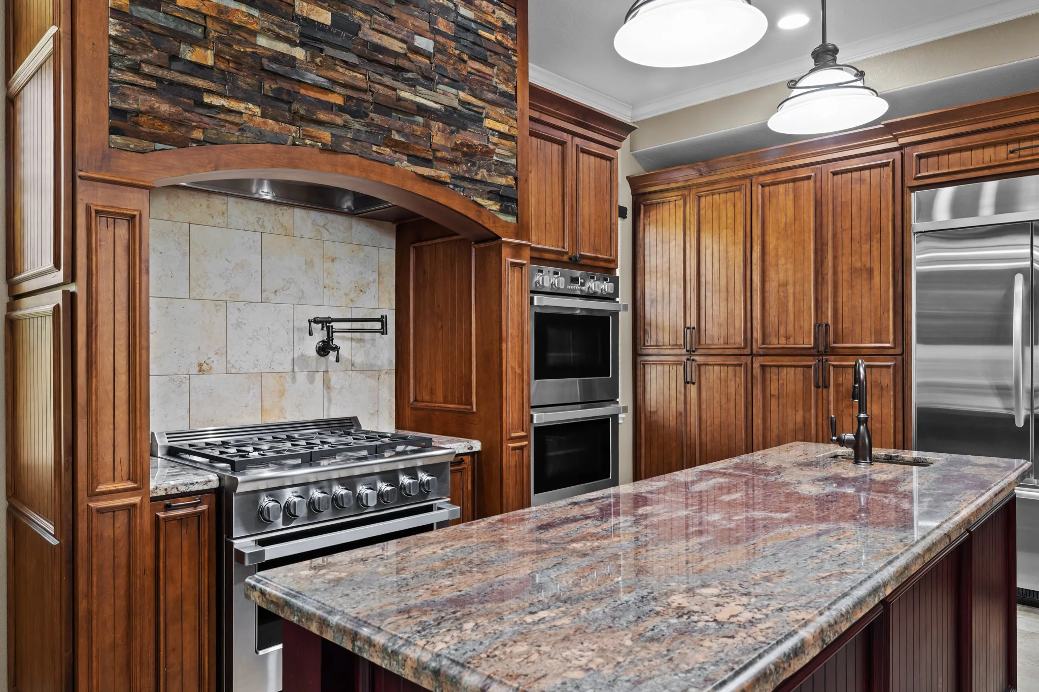 View of a kitchen with a granite countertop, wooden cabinets, a stainless steel oven, and a black faucet in the island